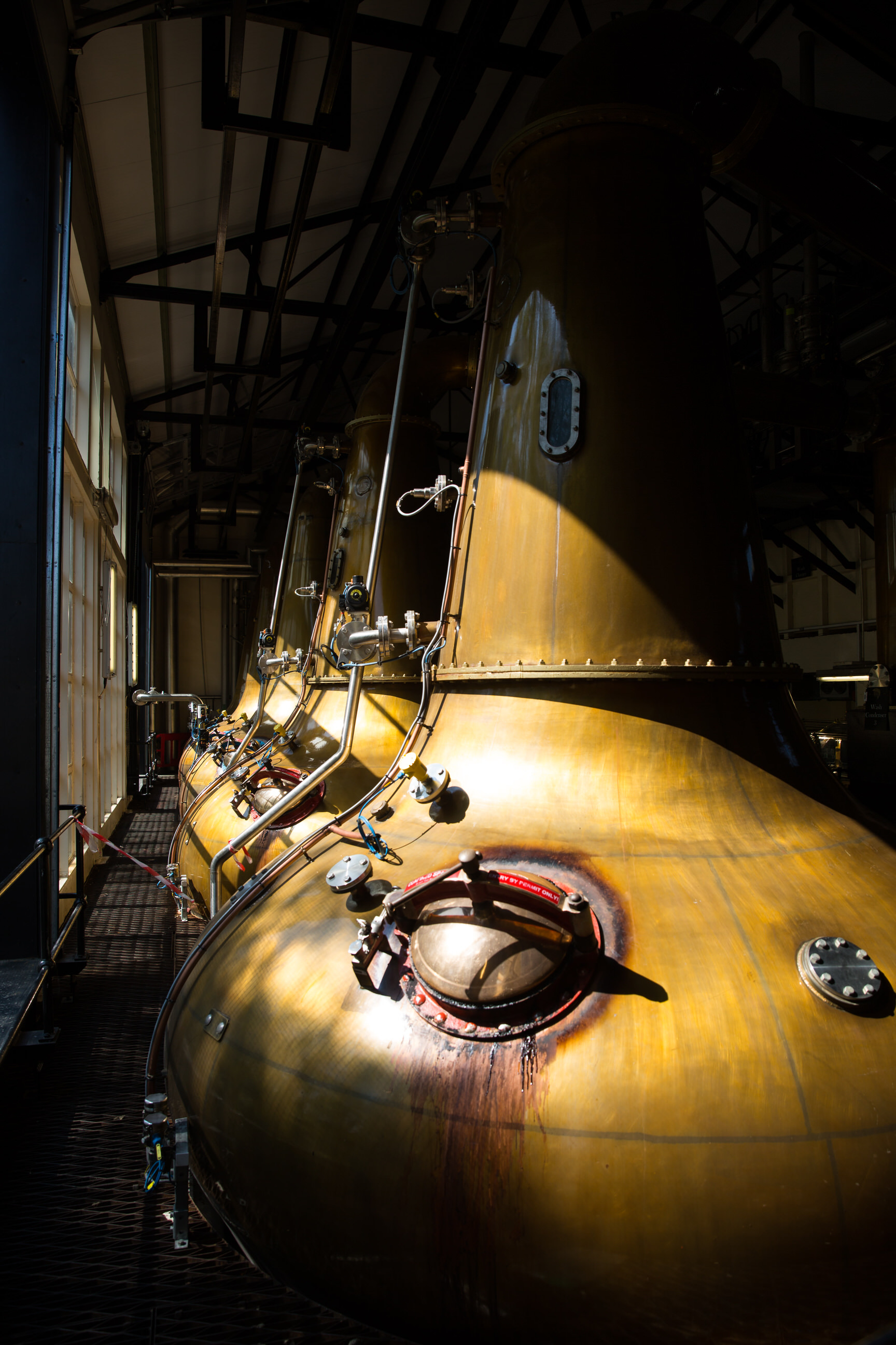 Large metallic cylindrical tanks inside an industrial facility or brewery, illuminated by natural light coming through windows, with various pipes and valves attached.