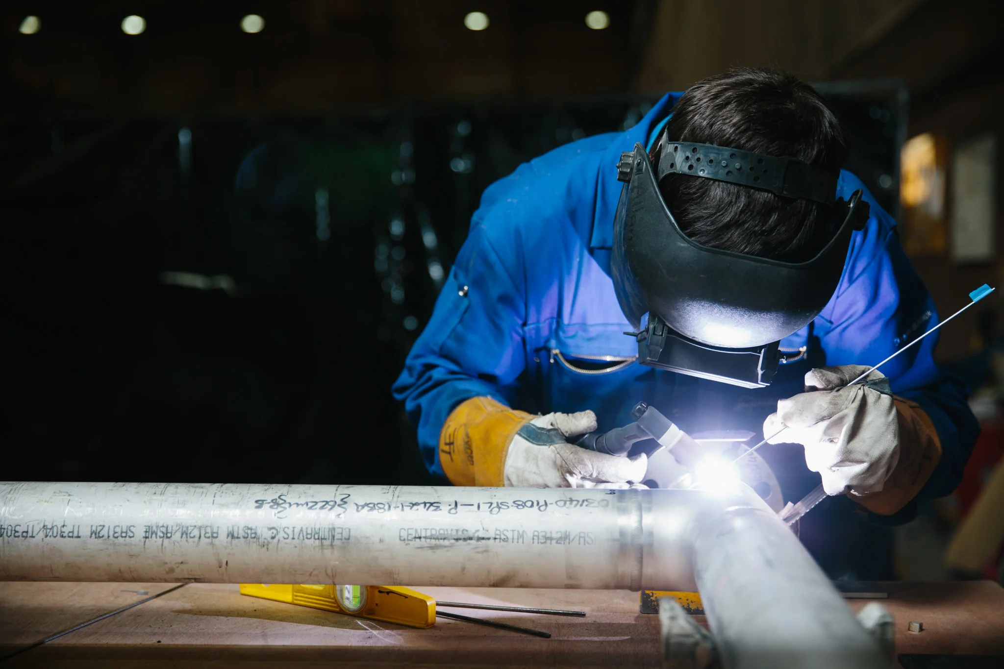 Worker wearing protective gear welding a pipe in an industrial setting.