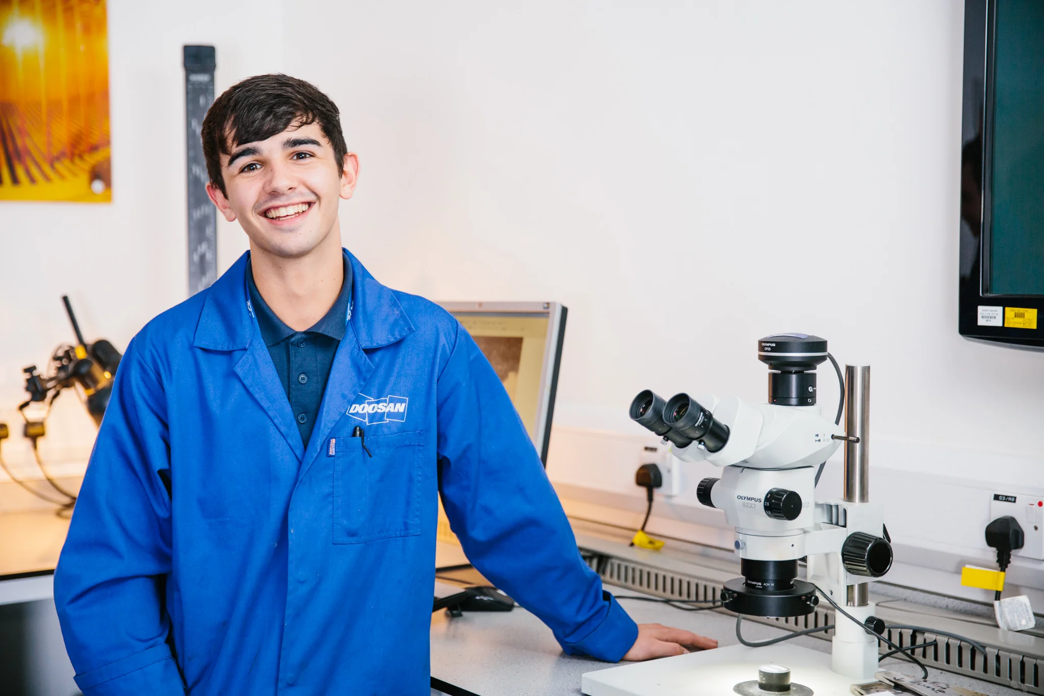 A young man with dark hair and a smile, wearing a blue lab coat with a Doosan logo, standing in a laboratory near a microscope and scientific equipment.