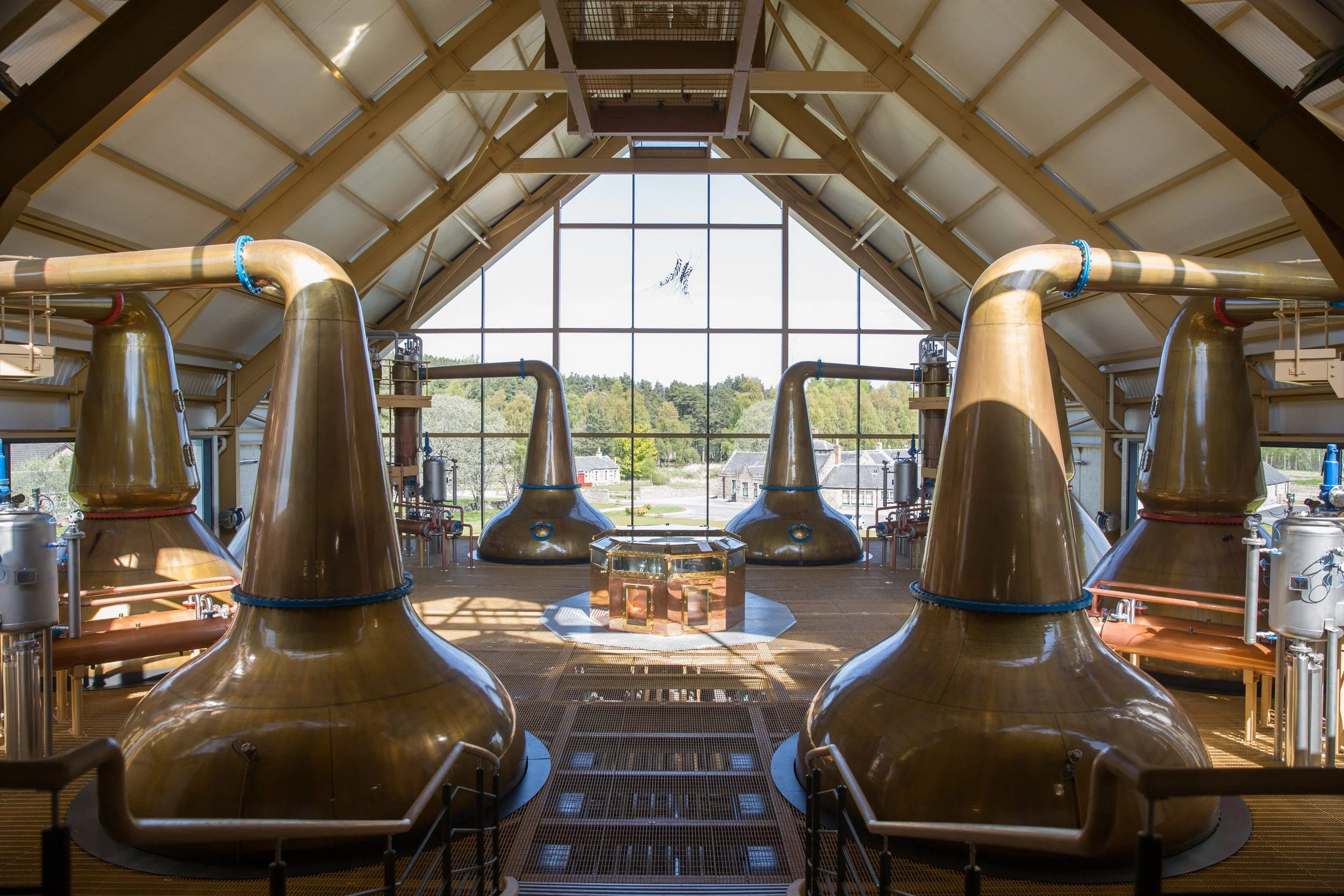 Interior of a large distillation or brewing facility with multiple copper stills and large piping, viewed from inside a glass-structured roof with rural landscape outside.