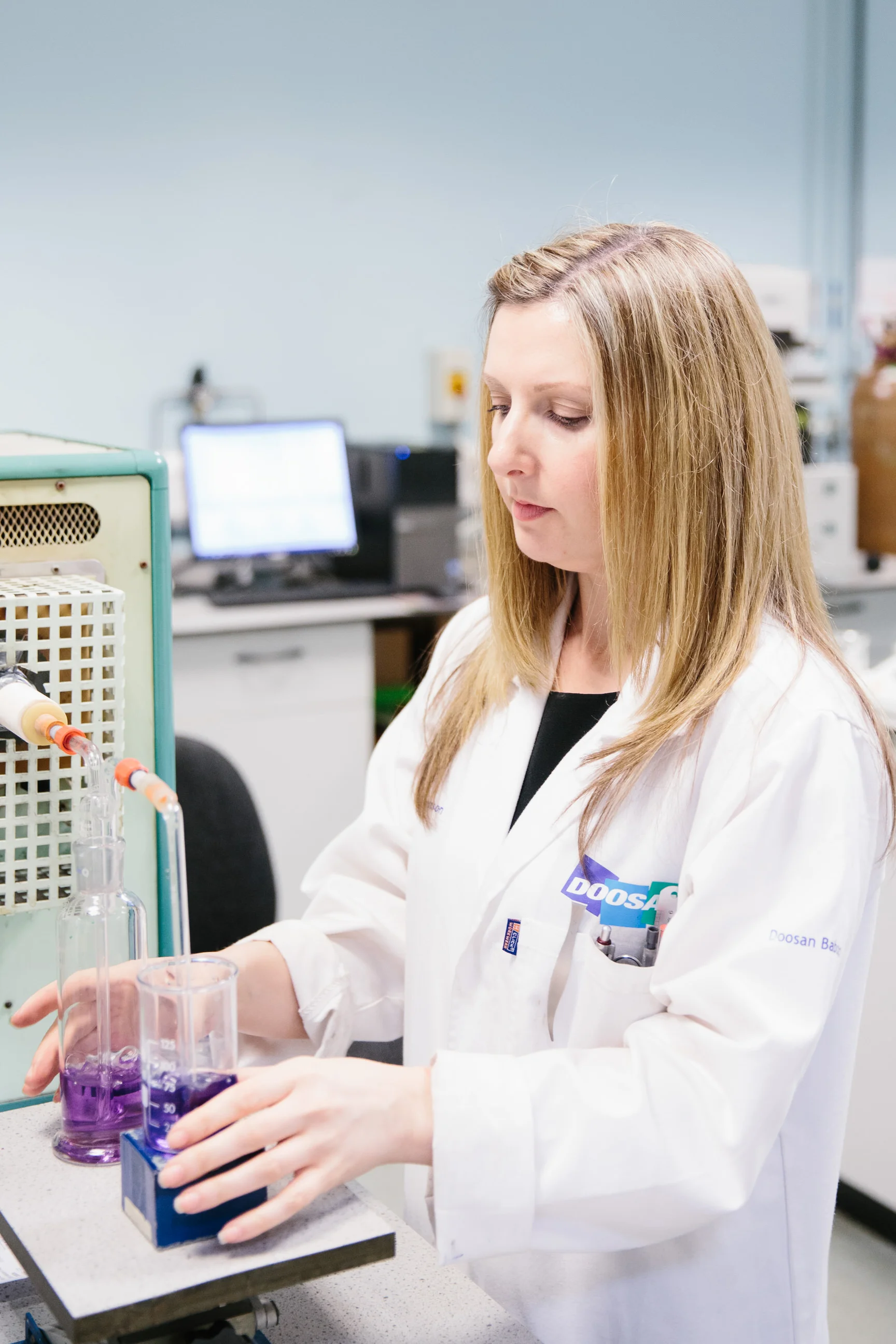 A woman in a white lab coat working with laboratory equipment, including beakers with purple liquid and glass tubes, in a science laboratory.