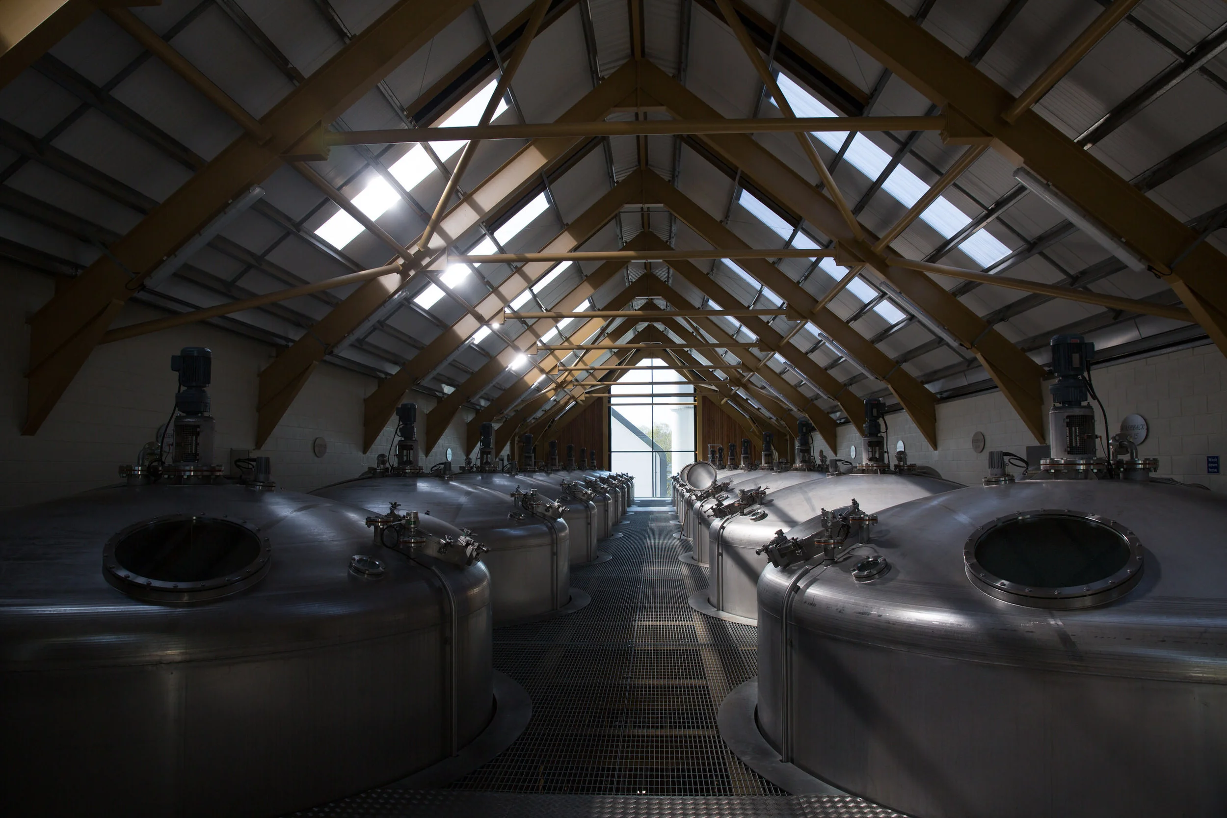 Interior view of a brewery or industrial facility with multiple large stainless steel tanks aligned in rows, wooden beams supporting the ceiling, and skylights allowing natural light to enter.