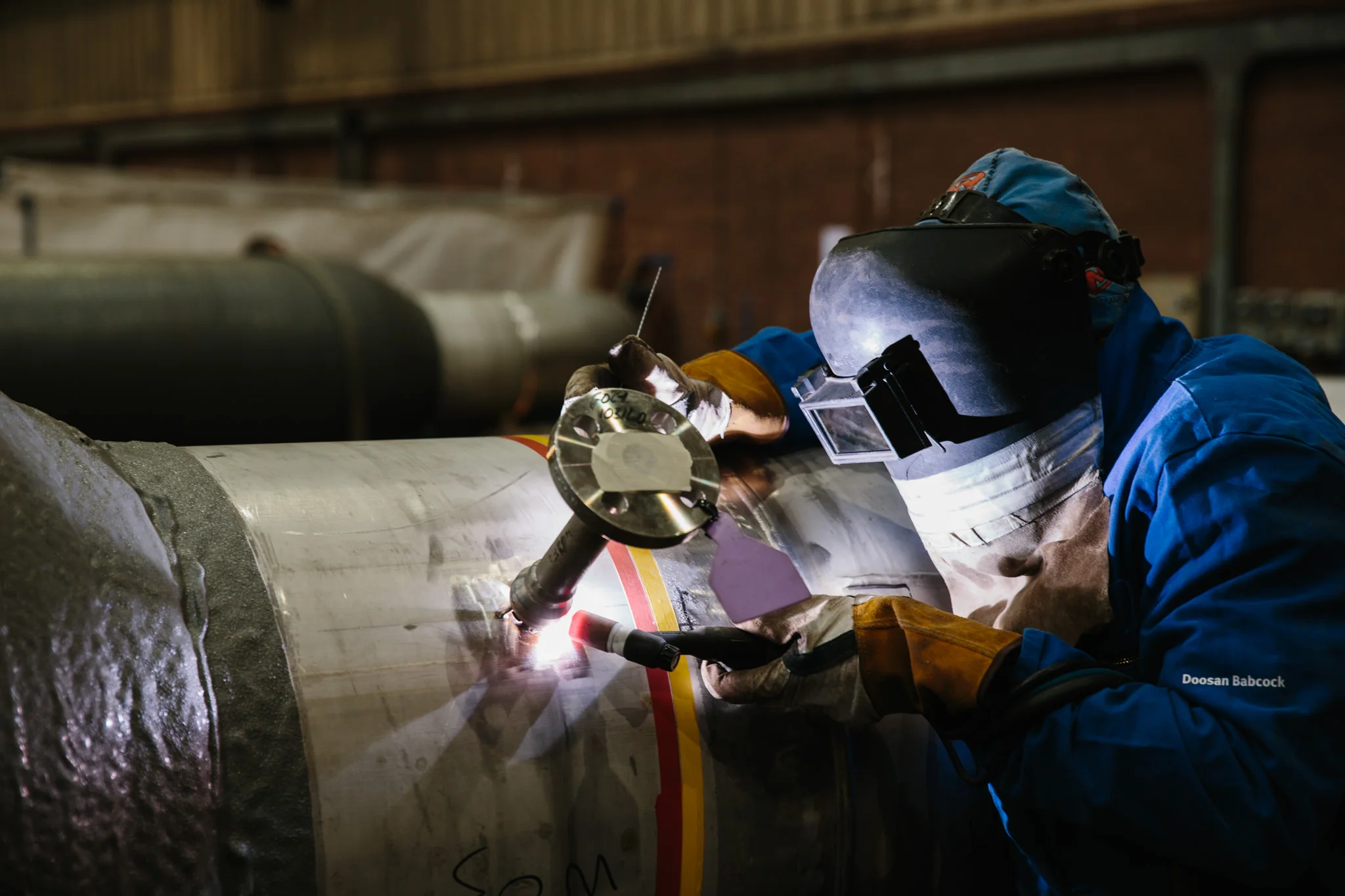 A worker wearing a welding helmet and blue protective jacket welding on a large metal pipe inside an industrial workshop.