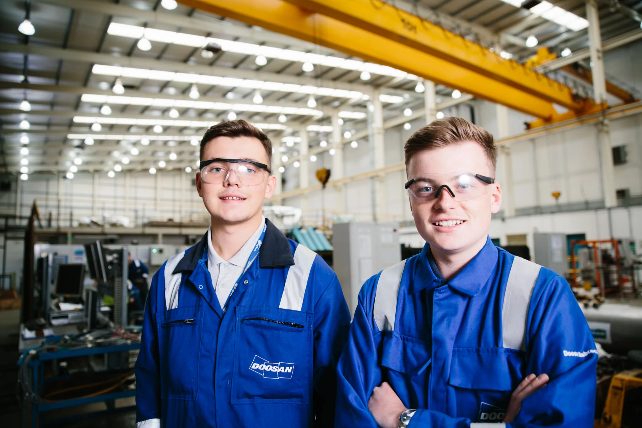 Two young men in blue uniforms with safety glasses standing inside an industrial warehouse with yellow overhead crane and industrial equipment.