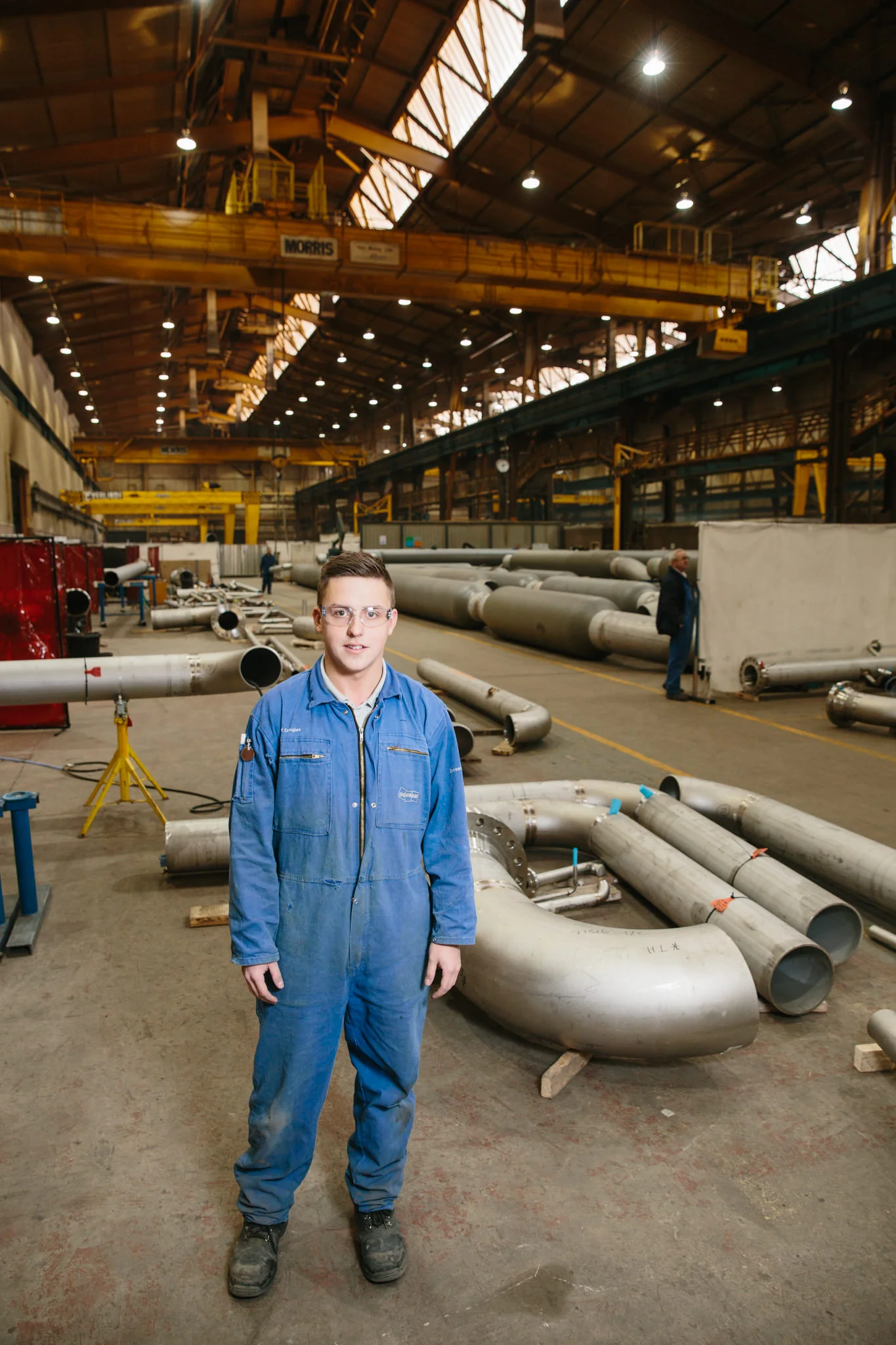 A young man in blue coveralls standing in an industrial warehouse with large pipes and equipment scattered around. The warehouse has high ceilings, metal beams, bright overhead lighting, and yellow overhead cranes.