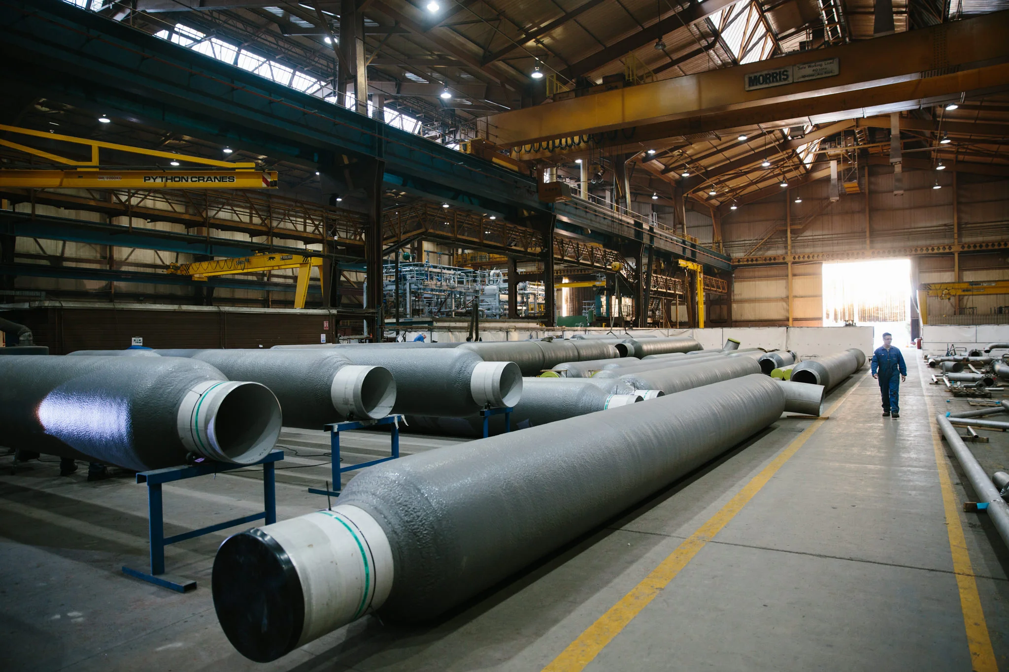 A spacious industrial warehouse with large pipes laid out on the floor, overhead cranes, and a worker walking towards the back. The warehouse has a high ceiling with skylights and is brightly lit with artificial lights.