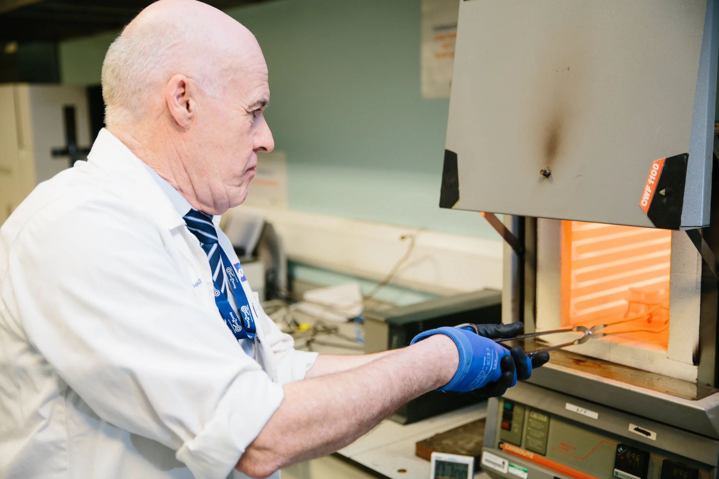 A man wearing a white lab coat, blue gloves, and a necktie working with a heating element and laboratory equipment in a laboratory setting.