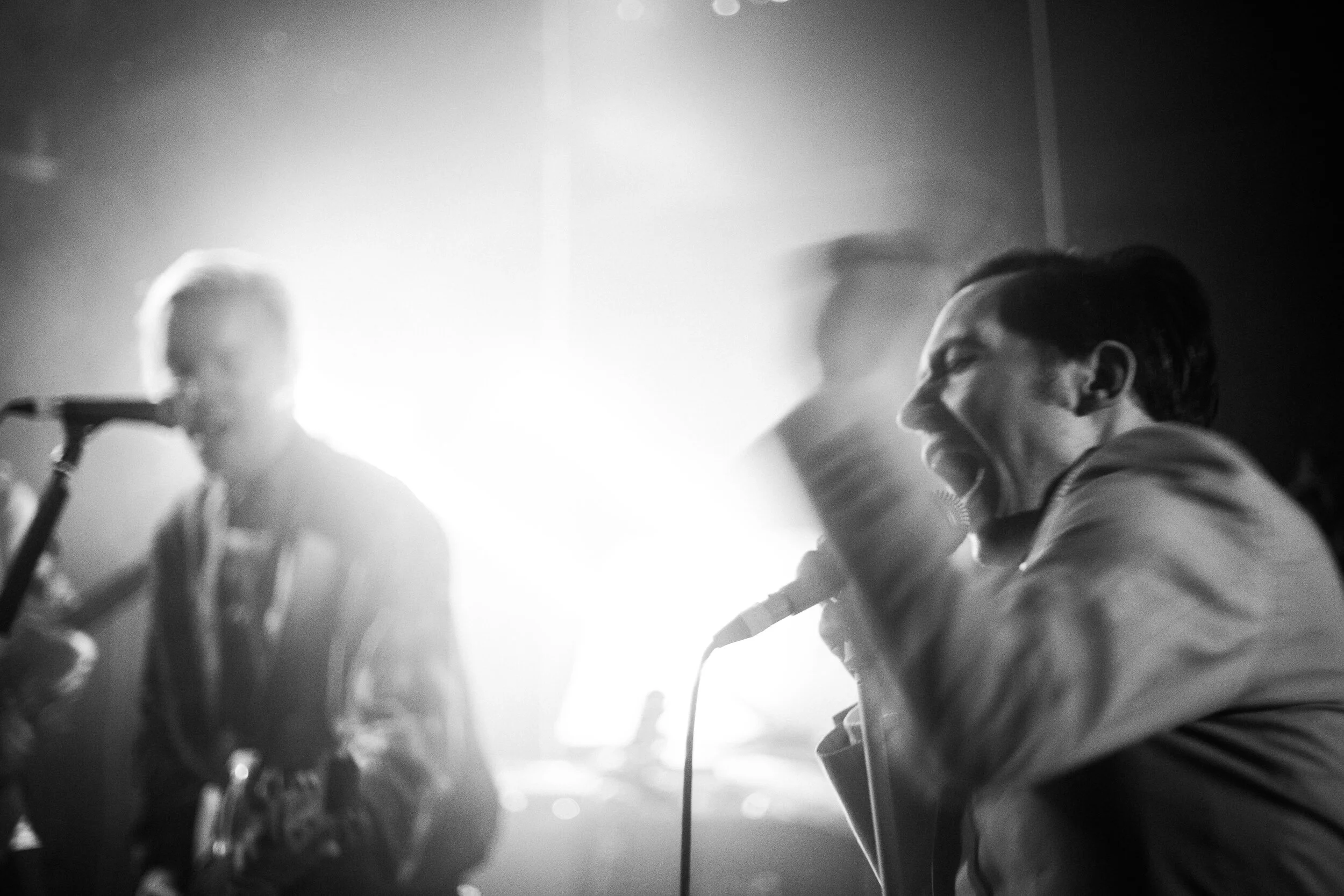 Black and white photo of a band performing on stage with a singer and a guitarist in focus, backlit creating silhouettes.