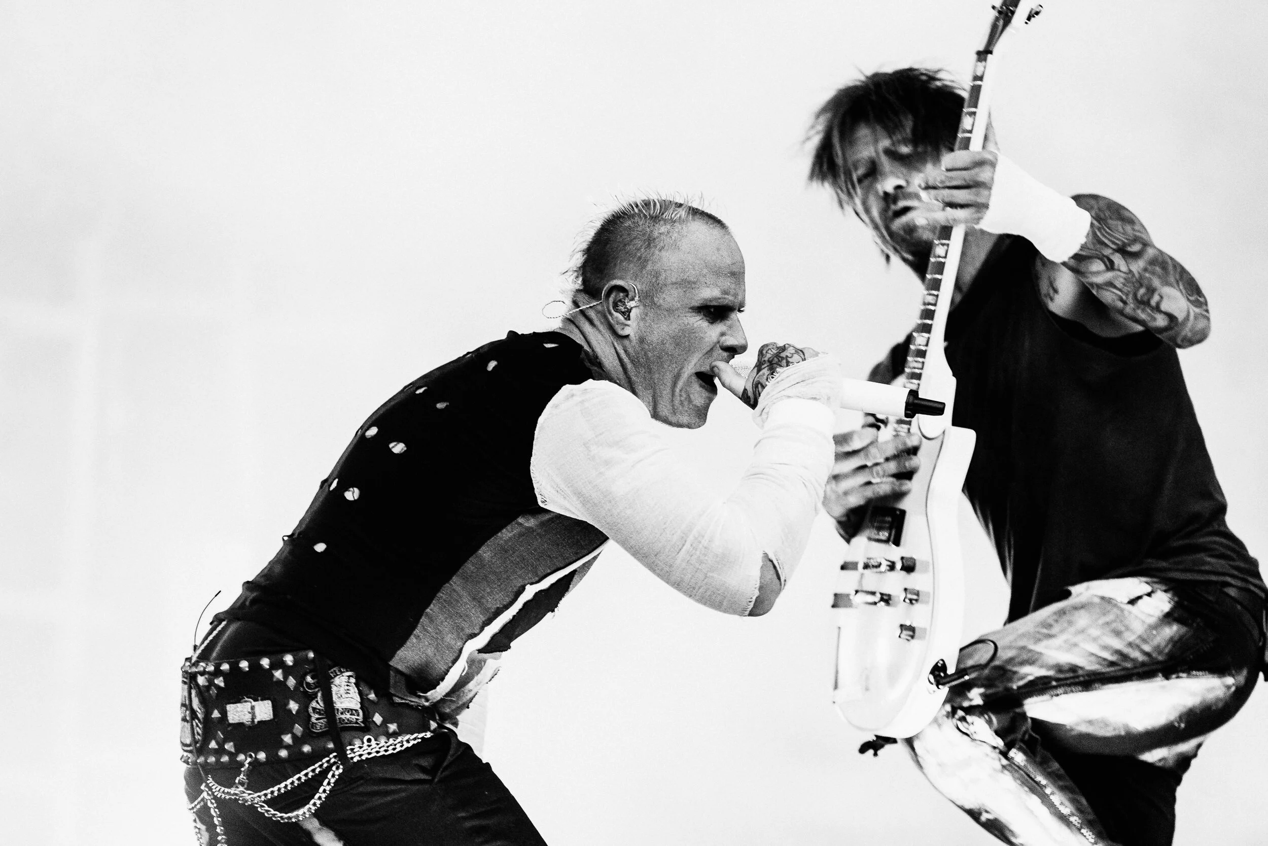 Two musicians performing on stage, one playing a guitar and the other singing into a microphone, both with tattoos and wearing dark clothing.
