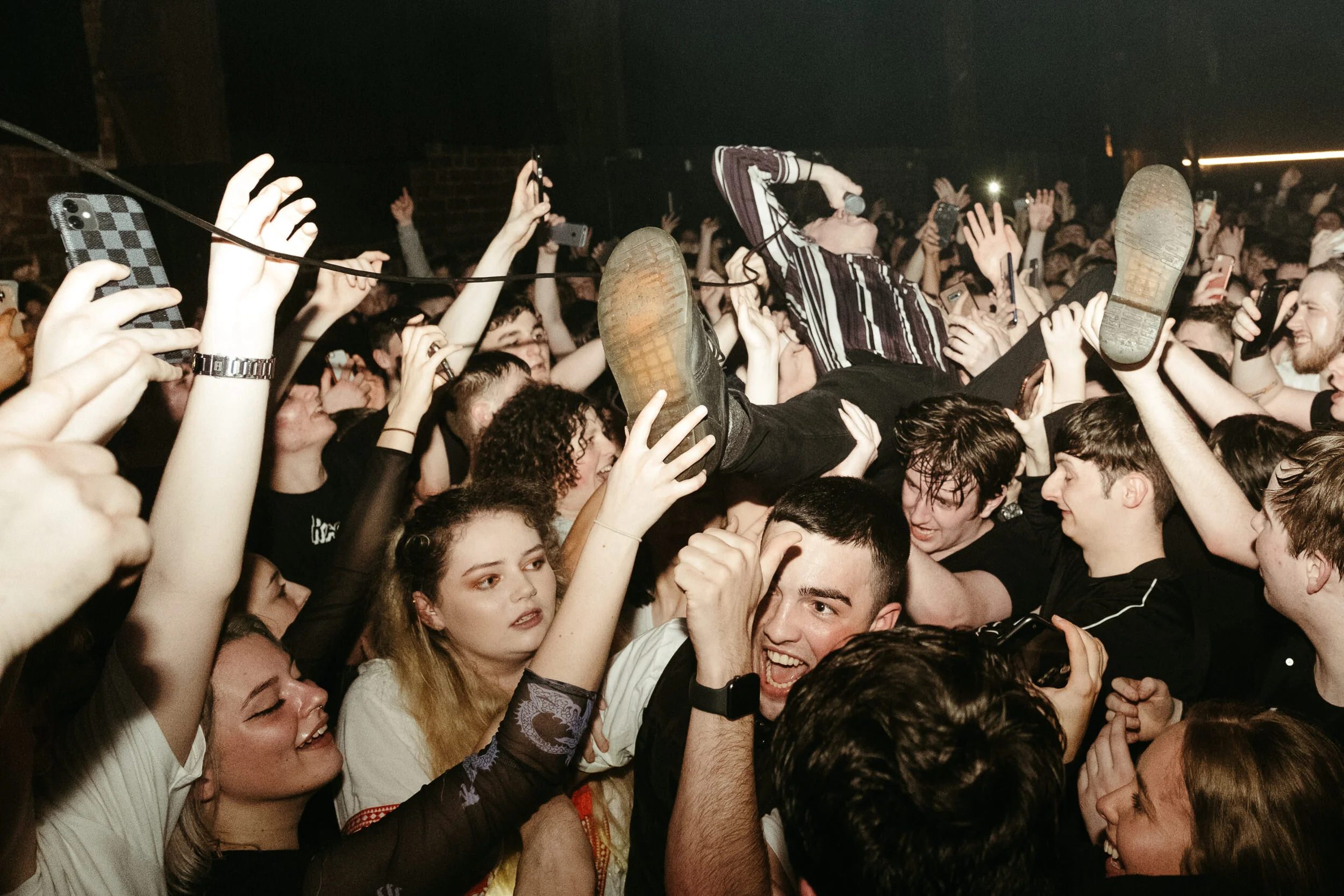A crowded concert scene with many people raising their hands and phones, a performer crowd surfing holding a microphone, and a lively atmosphere.
