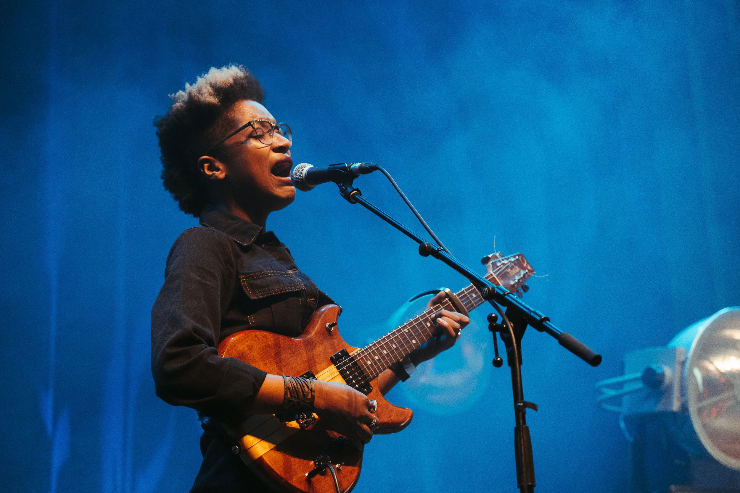 A woman singing into a microphone while playing an acoustic guitar on stage with a blue background.