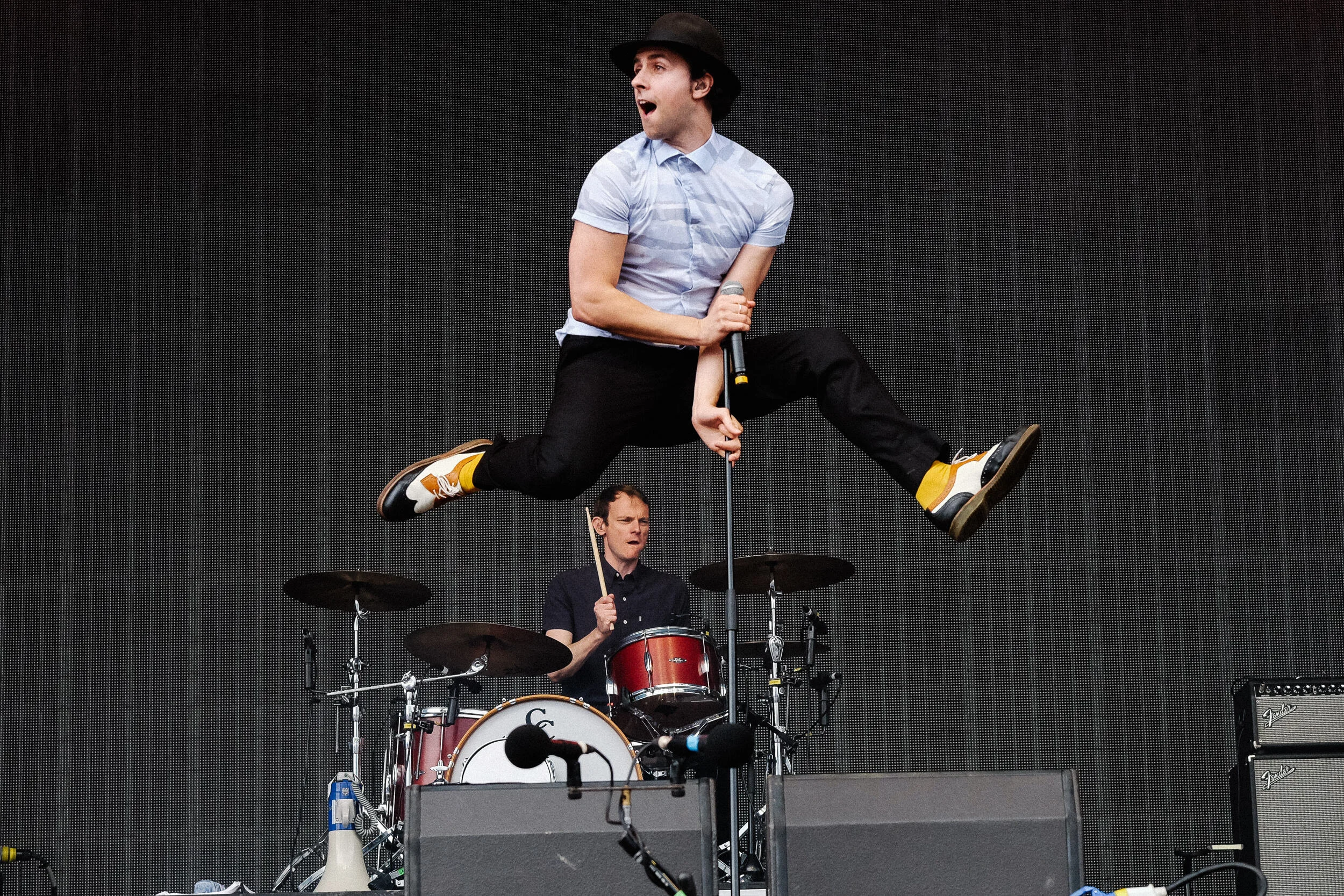 A musician wearing a hat and light blue shirt jumps in the air while performing with a microphone during a live concert. A drummer in the background plays a red drum set.