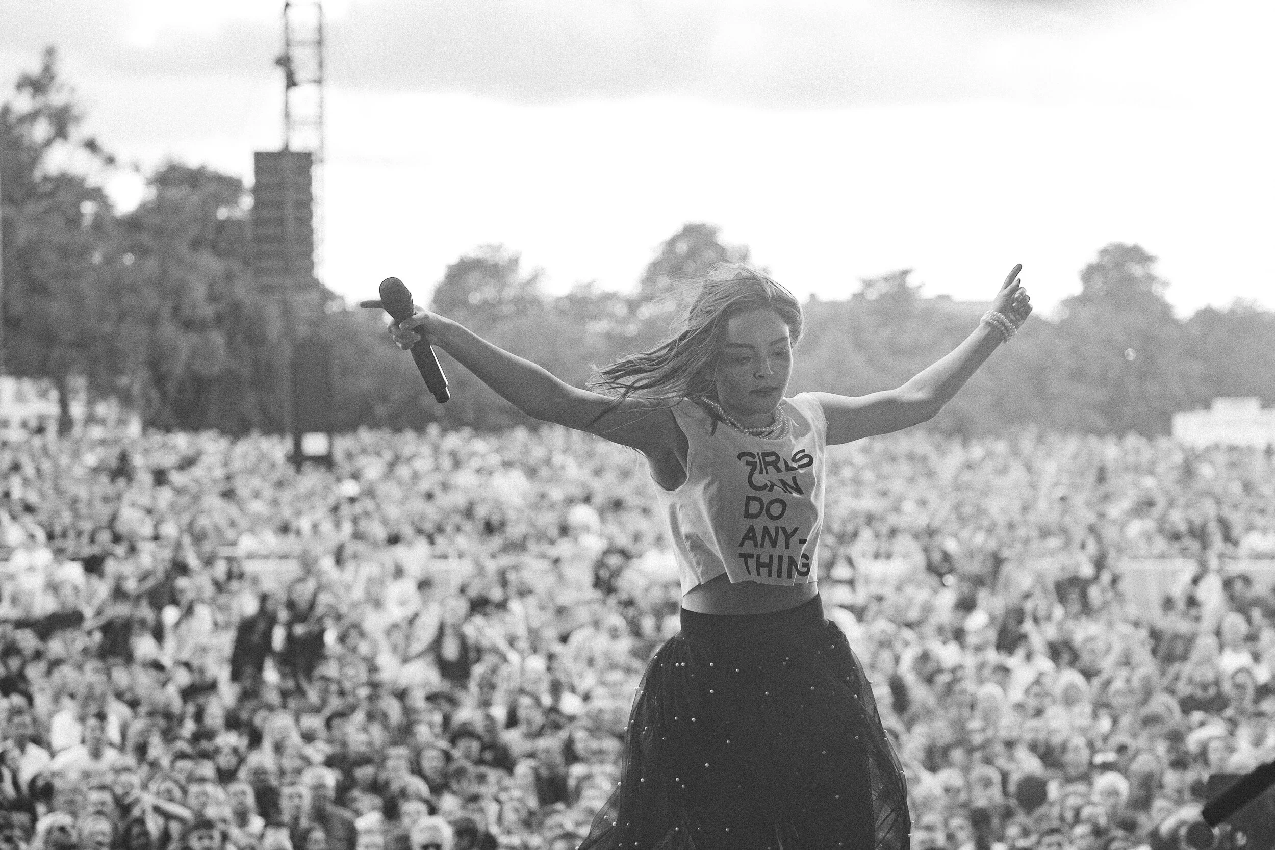 A young woman with long hair holding a microphone and dancing on stage during a concert, with a large crowd of people in the background.