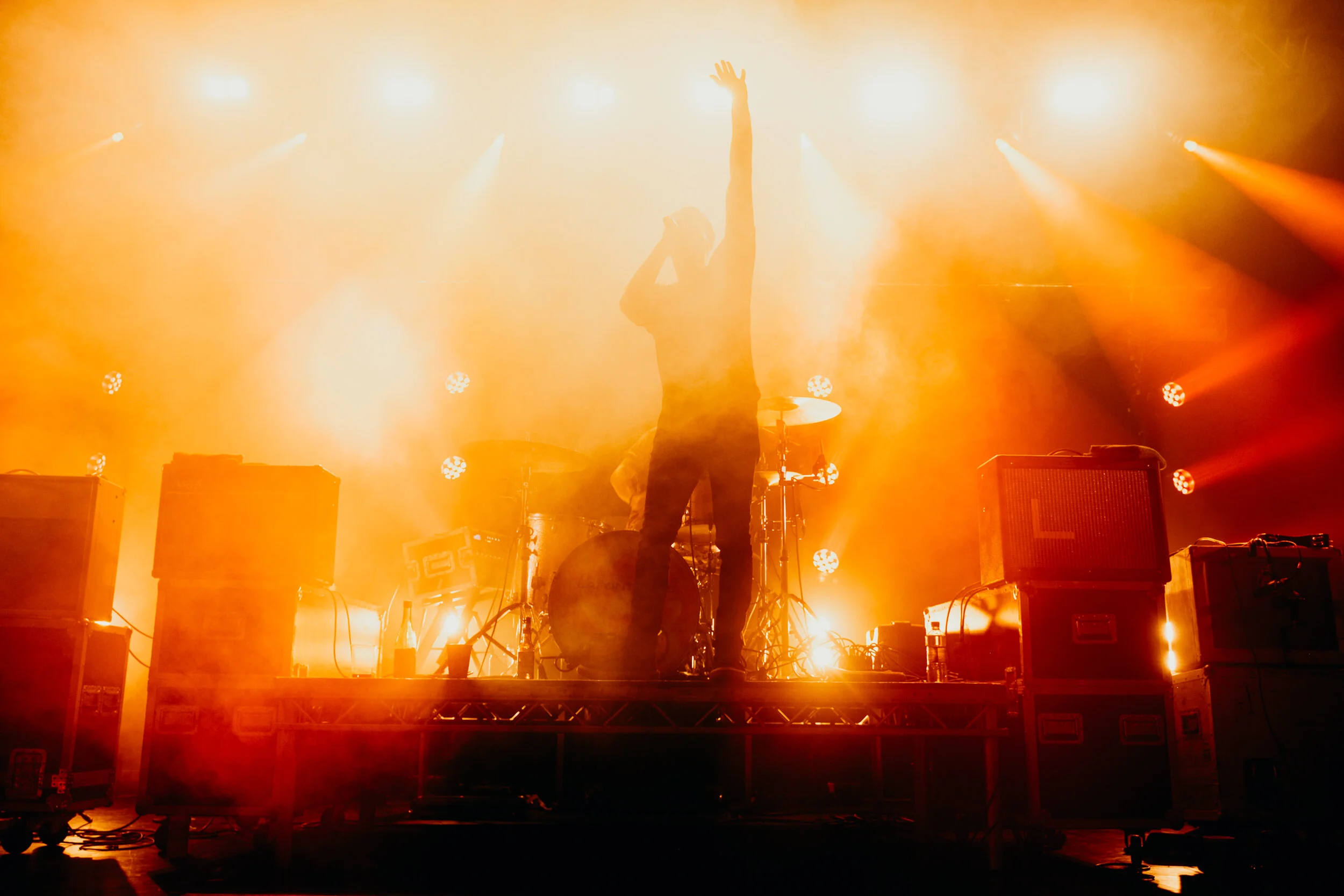 A singer performs on stage with bright orange lighting, standing with one arm raised and holding a microphone. Musical equipment and stage monitors are visible around them.