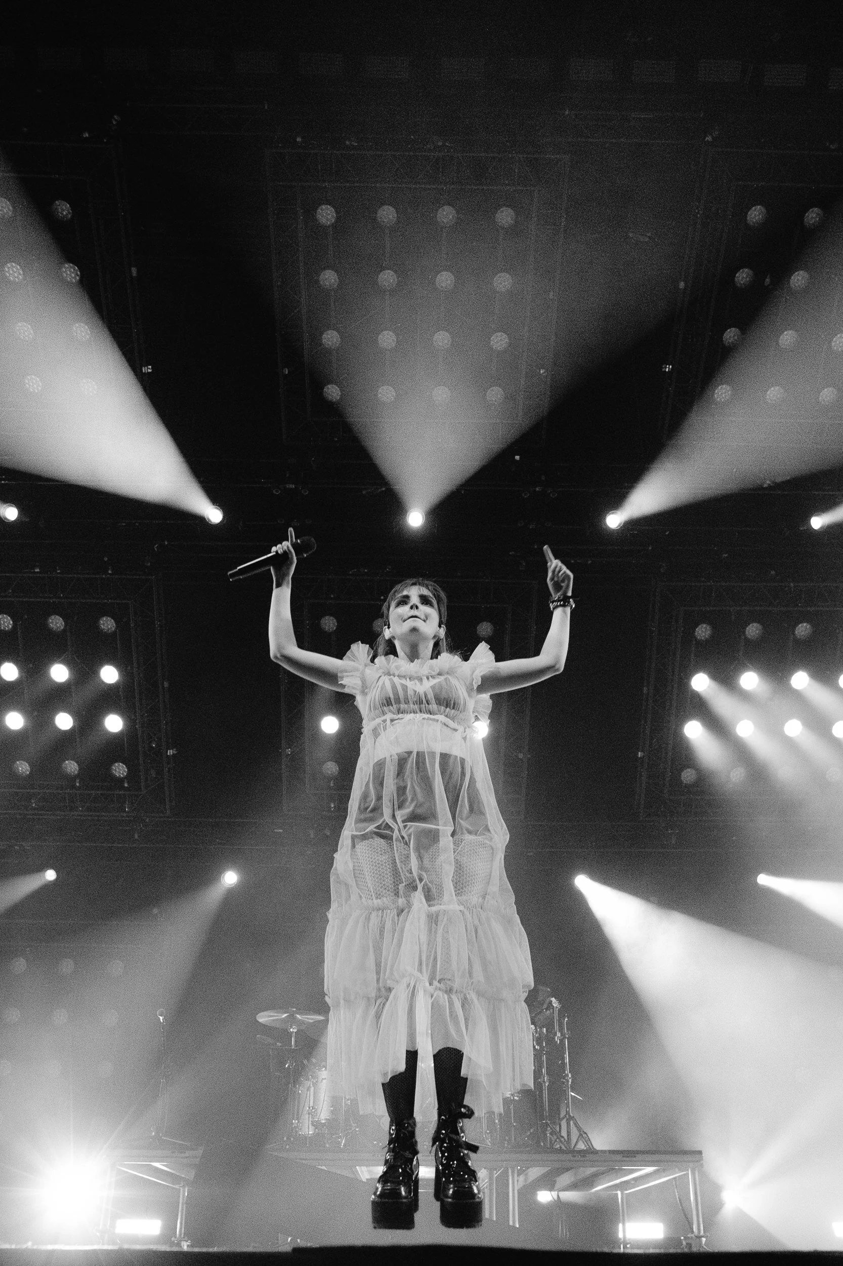 A female performer on stage holding a microphone in her right hand. She is wearing a flowing, layered dress and black combat boots. Stage lights illuminate her from behind, creating a dramatic effect.