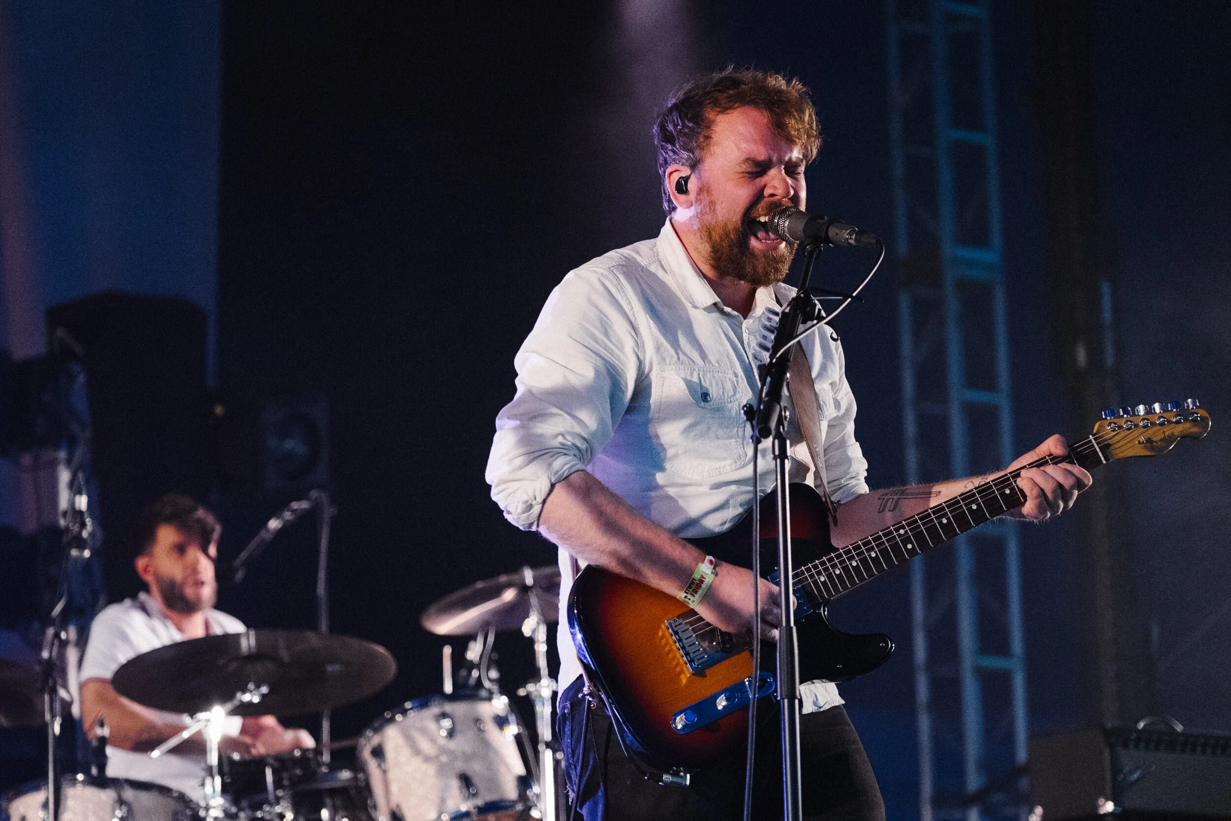 A musician passionately singing into a microphone while playing an acoustic guitar on stage, with a drummer and another band member visible in the background.