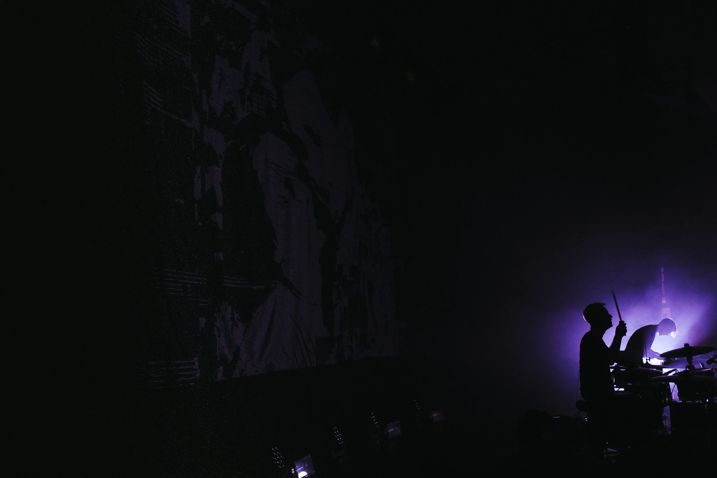 A dark scene with silhouettes of people, some playing musical instruments, illuminated by purple stage lighting.