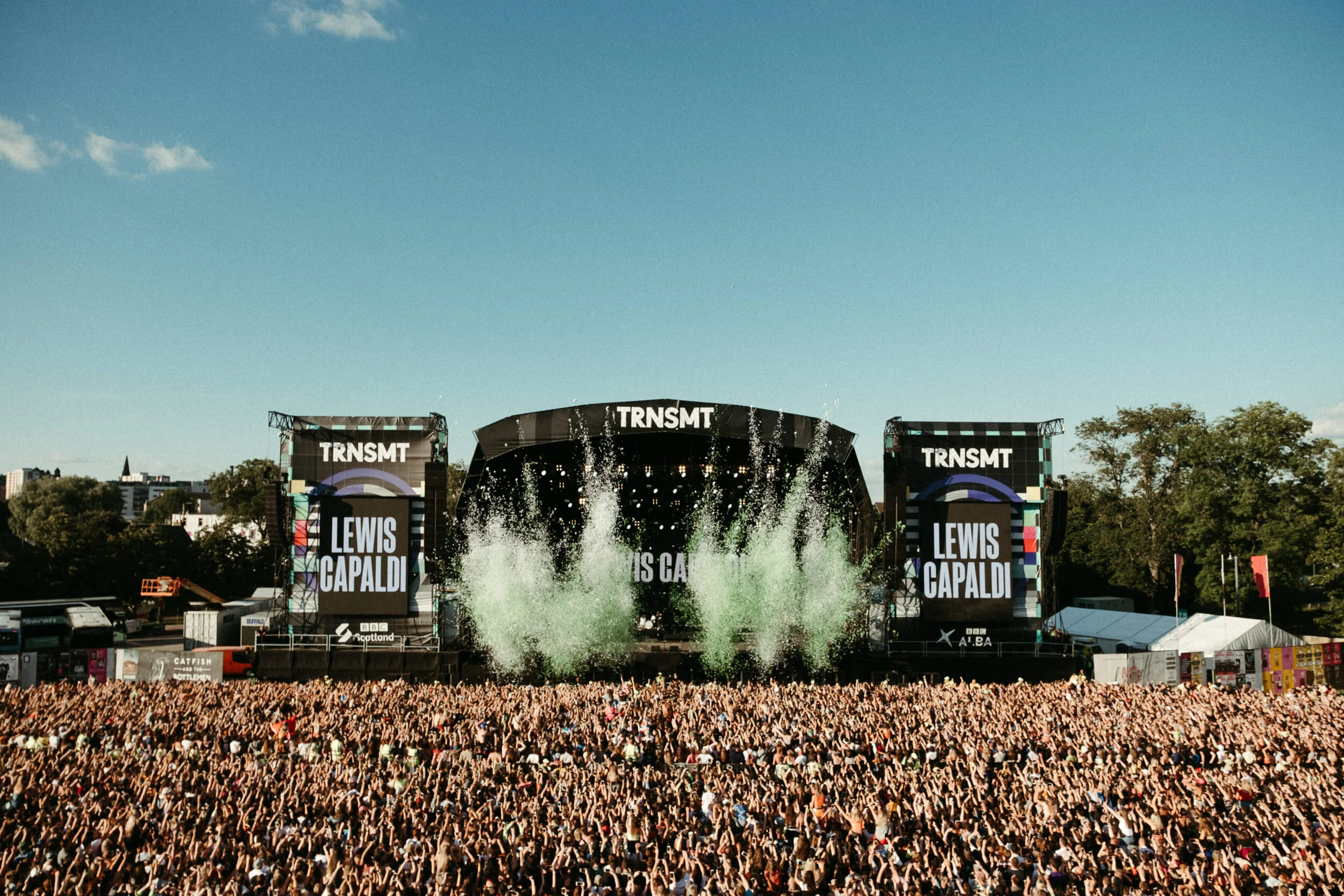 Large outdoor music festival with a stage displaying 'TRNSMT' and the name 'Lewis Capaldi' in front of a crowd of thousands of attendees, with trees and buildings in the background under a blue sky.
