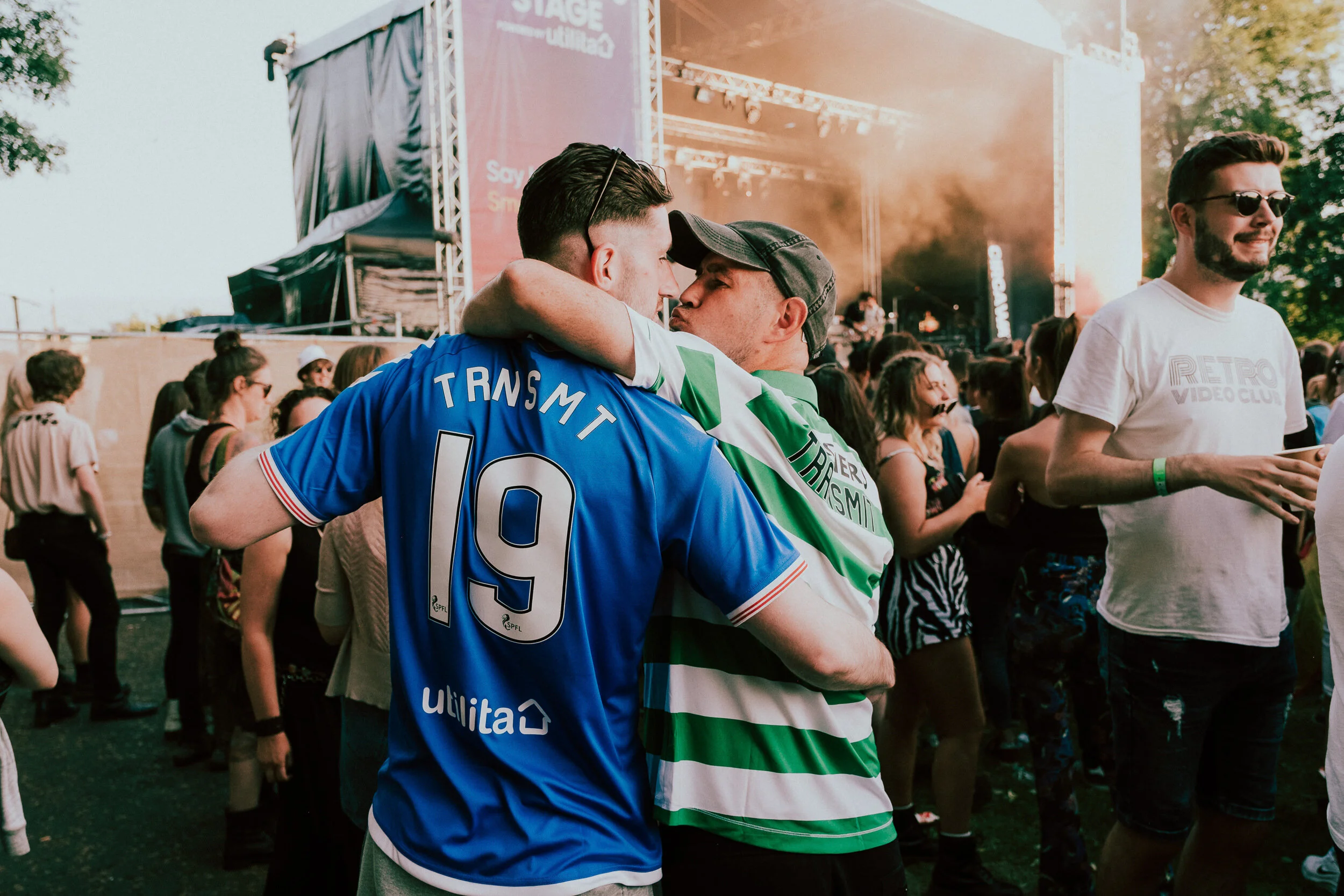 Two young men embrace each other at a concert or festival. One is wearing a blue sports jersey with number 9, and the other is wearing a white and green striped shirt. There is a stage with lighting and a crowd of people in the background, with some 