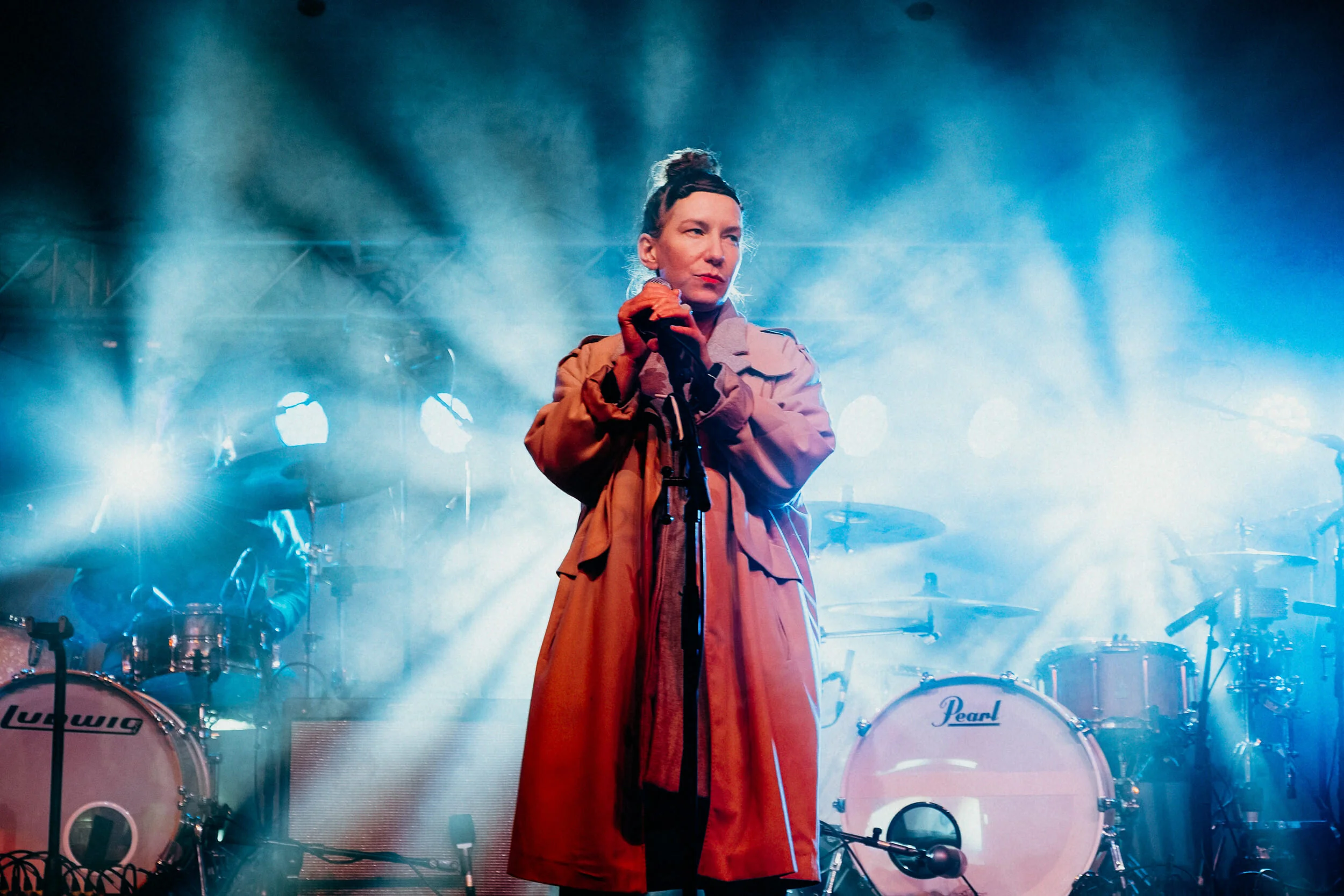 A female singer holding a microphone on stage with blue lighting, wearing a beige jacket and a red skirt, with drums and musical equipment in the background.