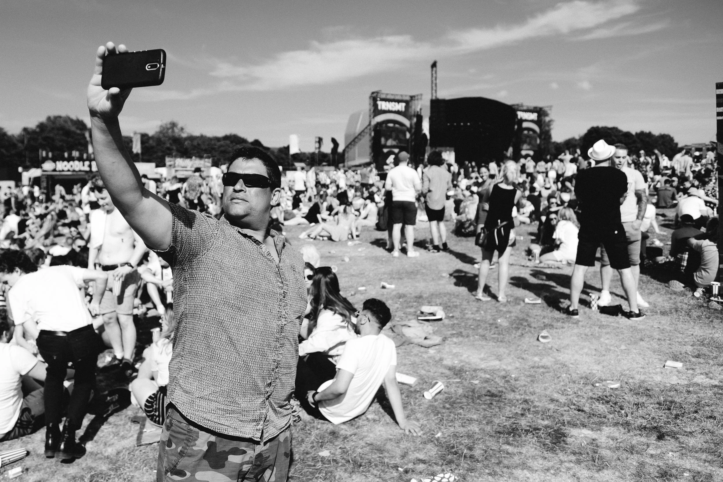 A man wearing sunglasses and a patterned shirt takes a selfie at an outdoor event with a large crowd gathered around. Many people are seated on the grass, and a stage with the word 'TRANSIT' is visible in the background.