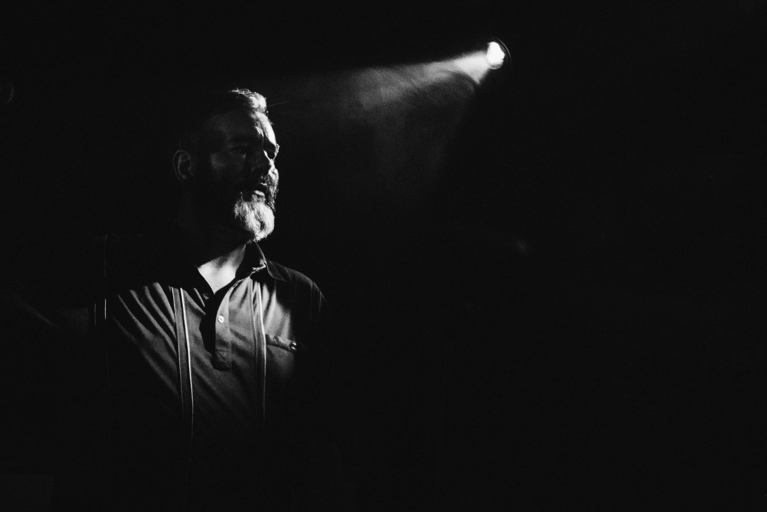 A man with a beard and mustache, wearing a dark jacket, is illuminated by a single light source in a dark environment.