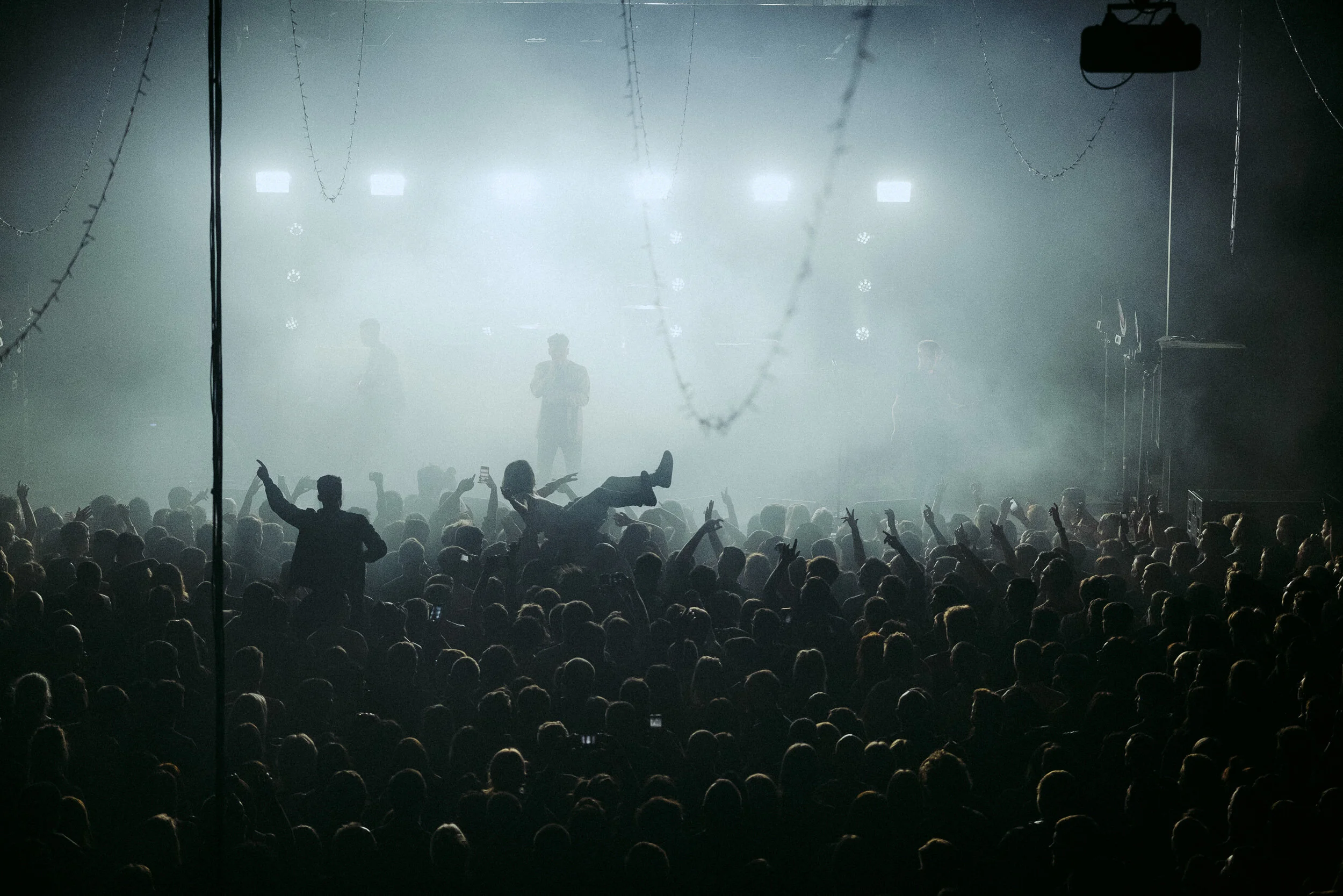 A large crowd attending a live concert or music event with a bright stage in the background and performers visible on stage. The audience is visible in the foreground, some raising hands, others capturing the moment with phones.