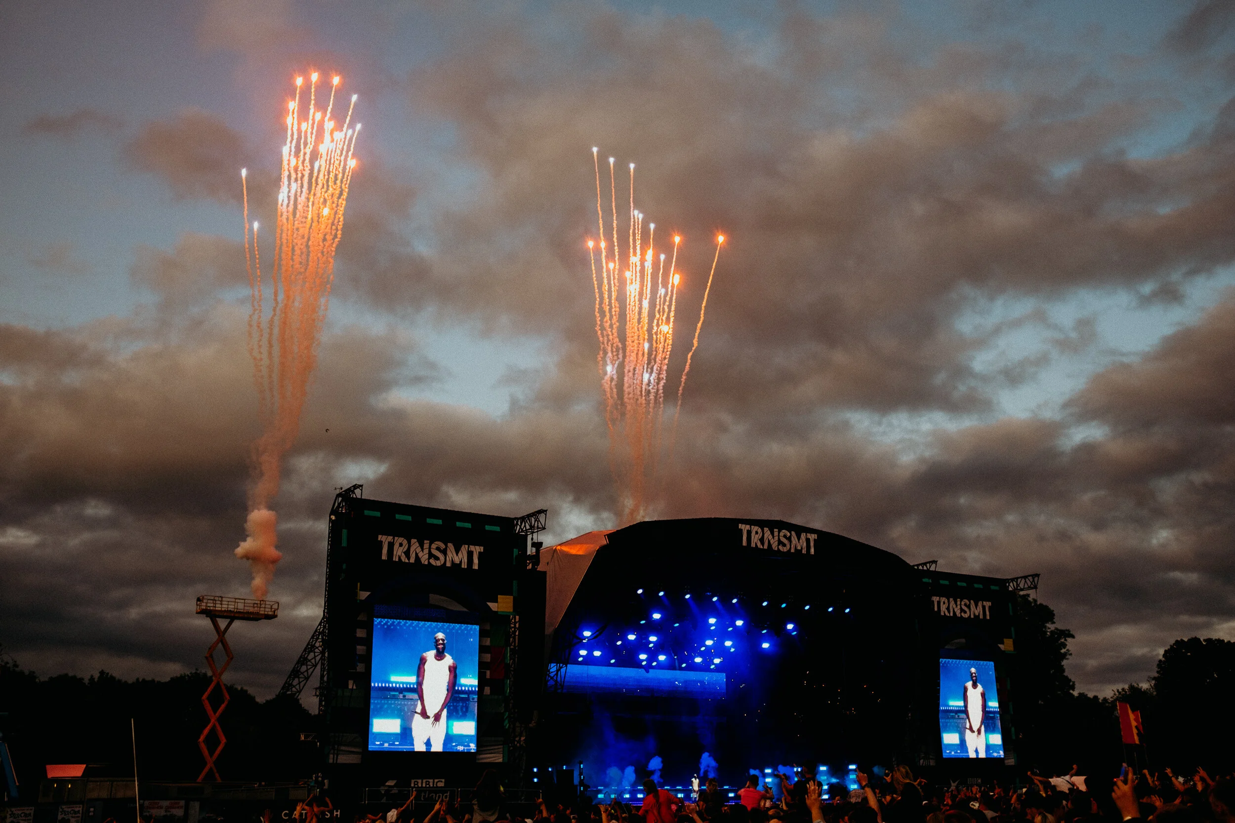 Fireworks light up the sky above an outdoor music festival stage with large screens displaying a performer. The crowd is visible in the foreground, enjoying the event.