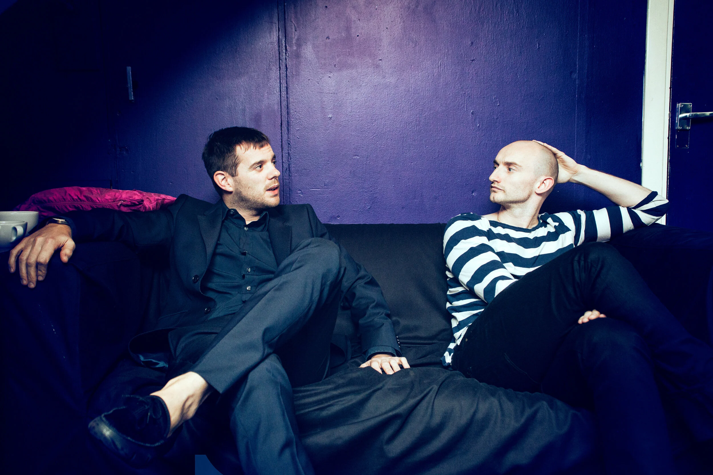 Two men sitting on a black couch against a purple wall. The man on the left has short dark hair and is dressed in a black suit, while the man on the right has a shaved head and is wearing a black and white striped shirt. The man on the right has one 