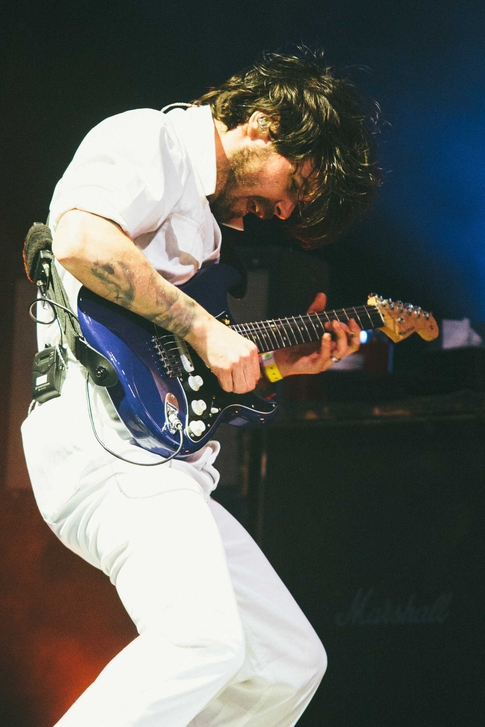 A male musician with long, dark hair playing an electric guitar on stage, wearing a white shirt and white pants.