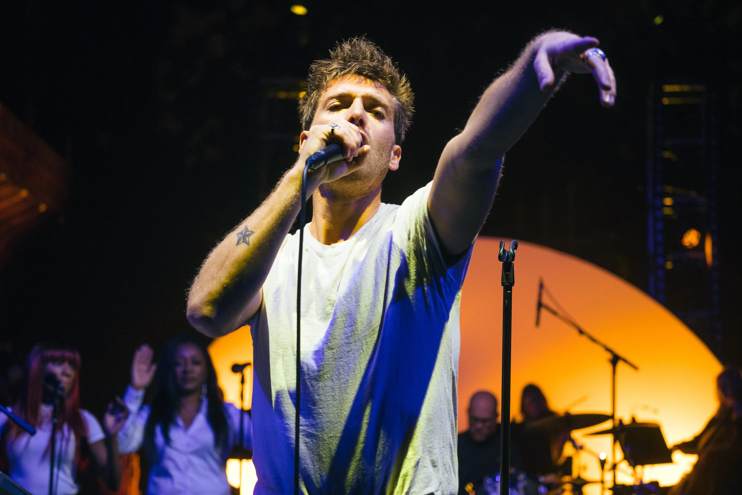 A male singer with short, spiked hair and tattoos, wearing a white t-shirt, performing on stage with a microphone, pointing outward, with audience members and stage lighting in the background.