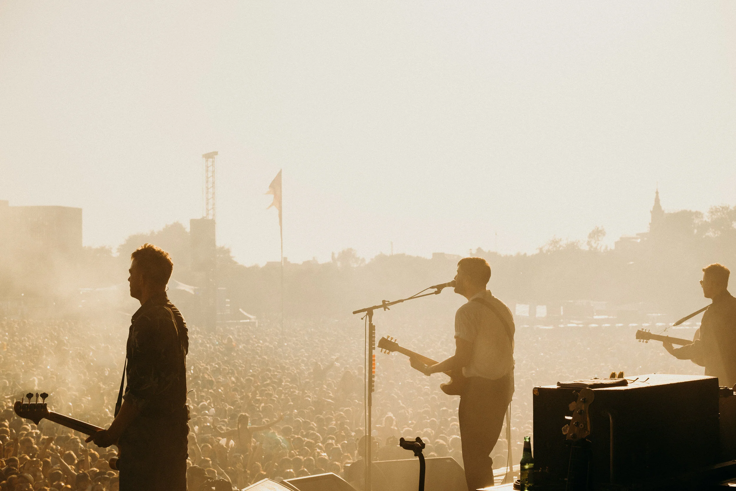 A live outdoor concert at sunrise with band members playing guitars and singing on stage in front of a large audience.