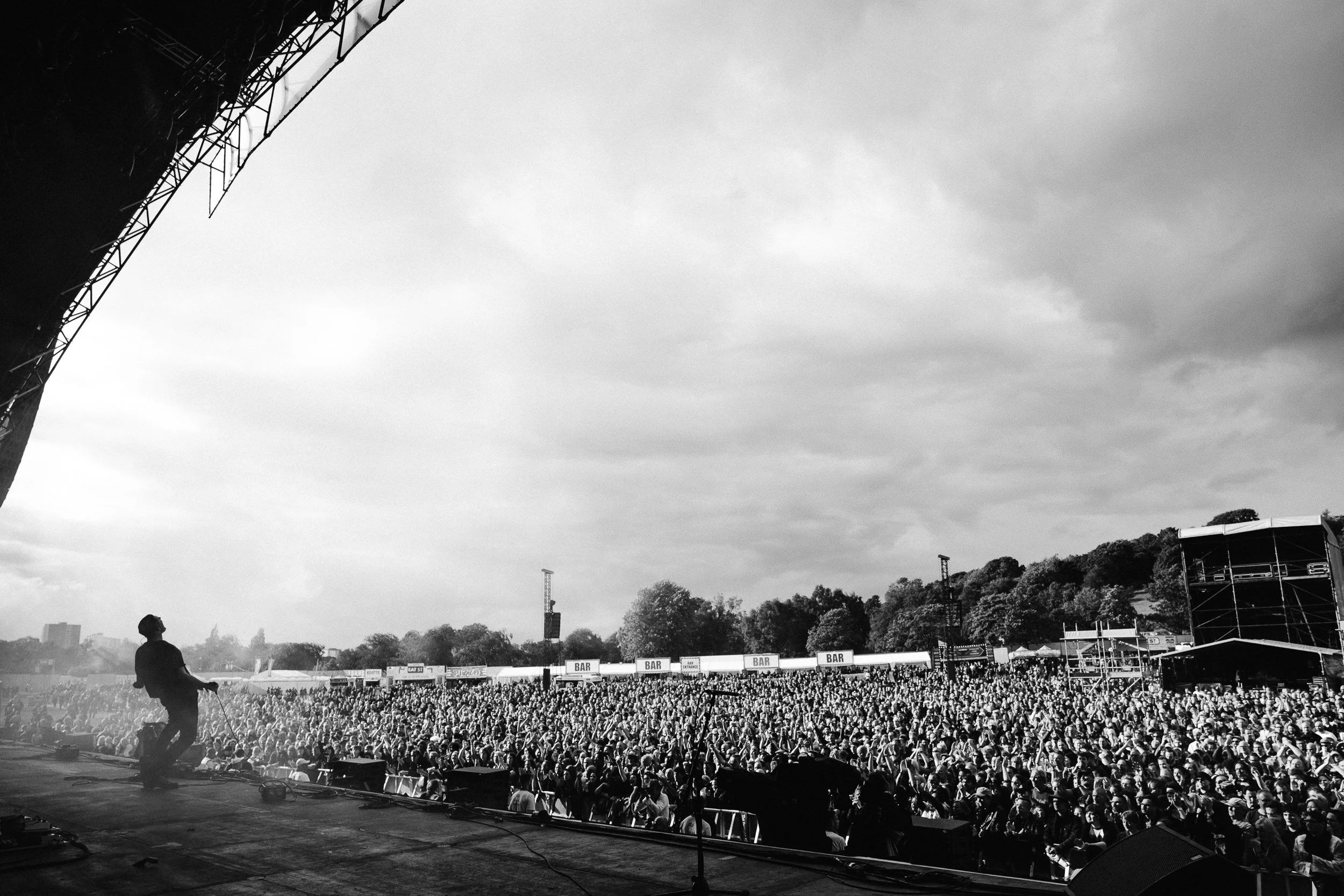 A large outdoor concert or festival with a stage and thousands of attendees; a person is standing on the stage looking at the crowd. The sky above is cloudy.