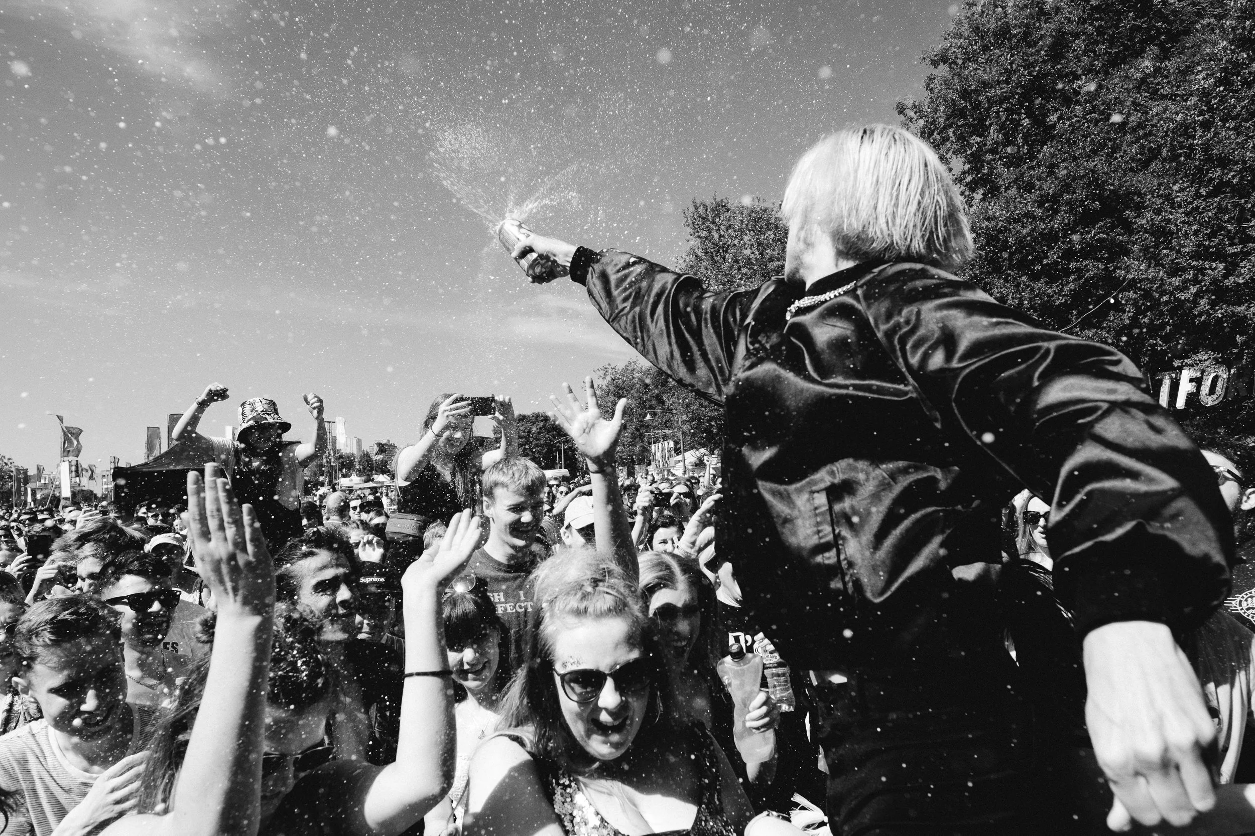 A large crowd of people gathered outdoors, celebrating with a person spraying a beverage into the air. The person has light-colored hair and is wearing a dark jacket. Many individuals are reaching out, smiling, and taking photos, with some raising th
