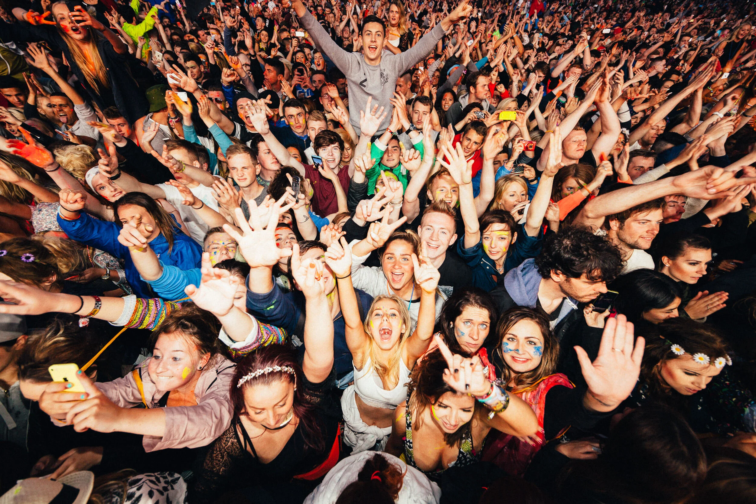 Crowd of people at a concert or festival with many smiling and raising their hands, some taking photos with smartphones, vibrant and energetic atmosphere.