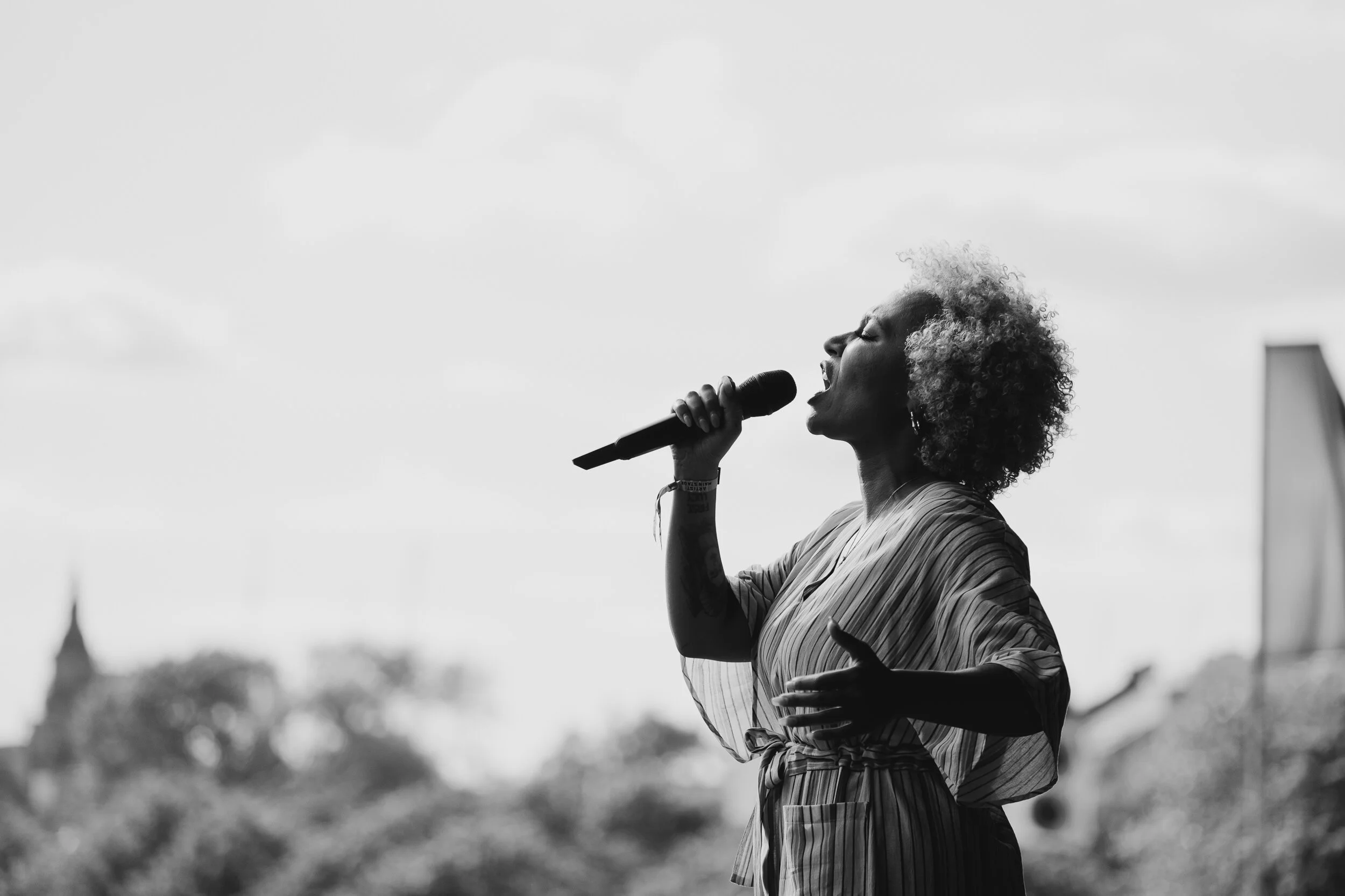 A woman with curly hair singing into a microphone outdoors on a cloudy day.