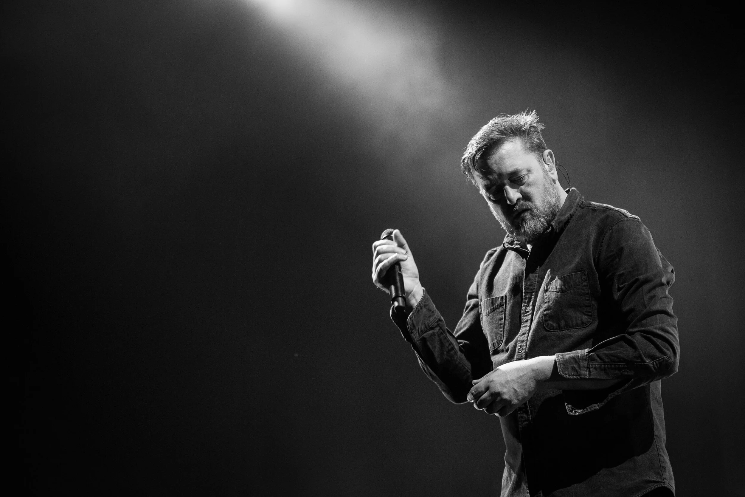 A black and white photo of a man with a beard and styled hair holding a microphone, looking down, standing on stage with a spotlight and dark background.