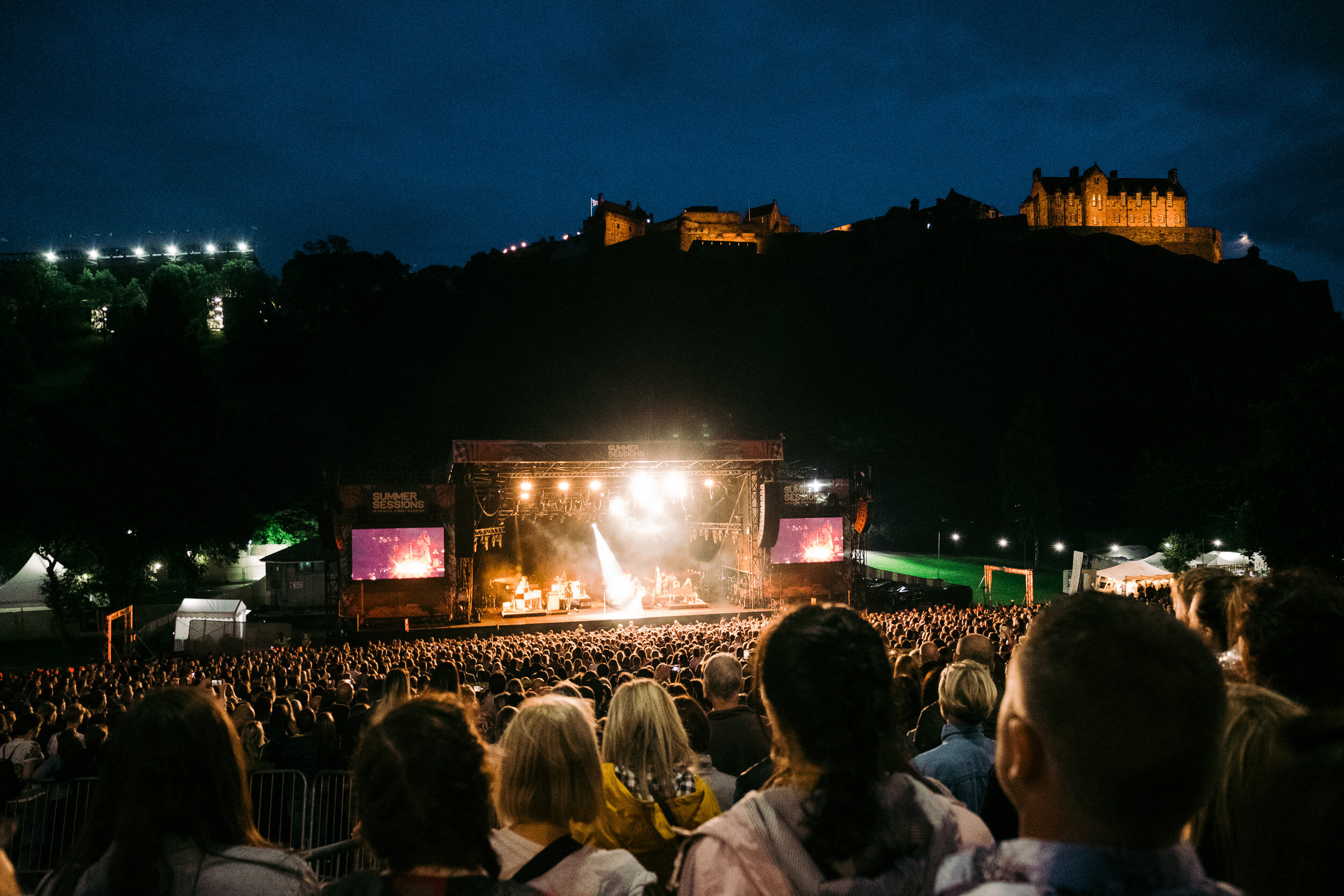 A large crowd of people attending an outdoor concert at night, with a stage illuminated with lights and screens, and a historic castle on a hill in the background.
