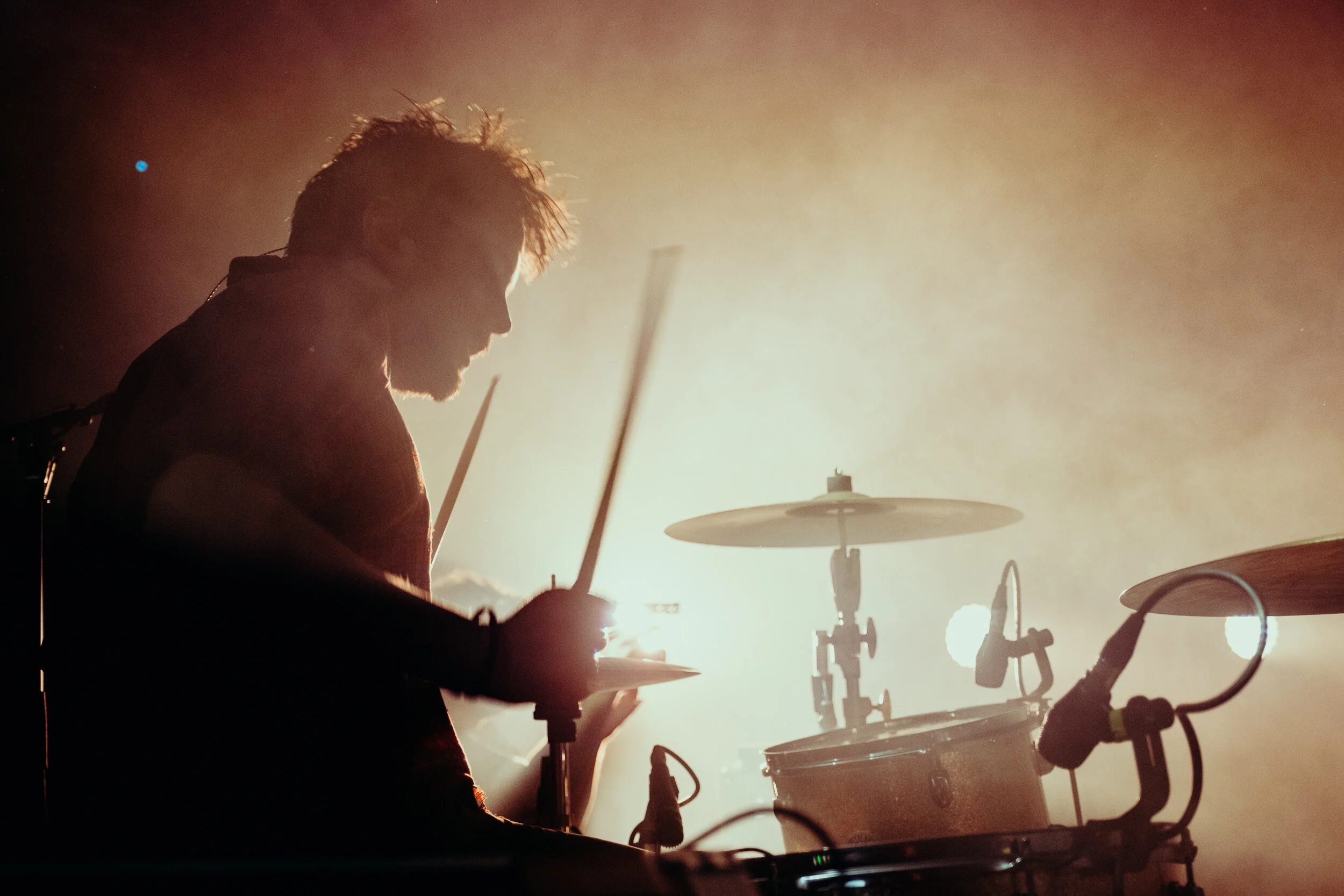 A silhouette of a drummer playing drums with a bright, hazy light in the background.