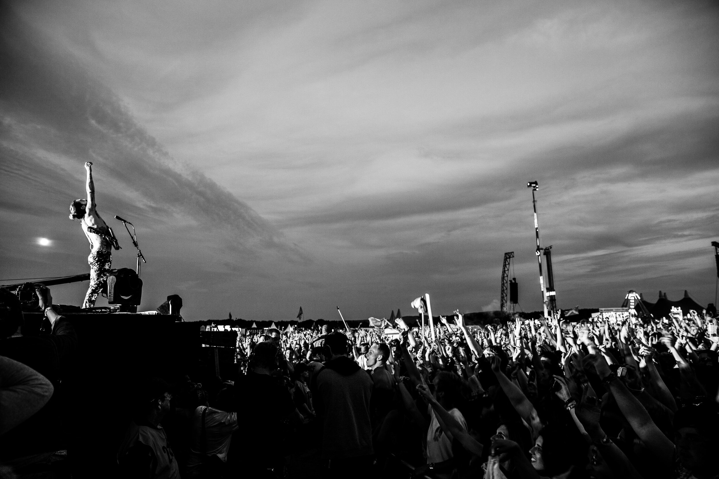 A musician on stage at a large outdoor concert, raising a fist to the crowd with a microphone in hand, while the audience has hands raised and cheers. The scene is in black and white.