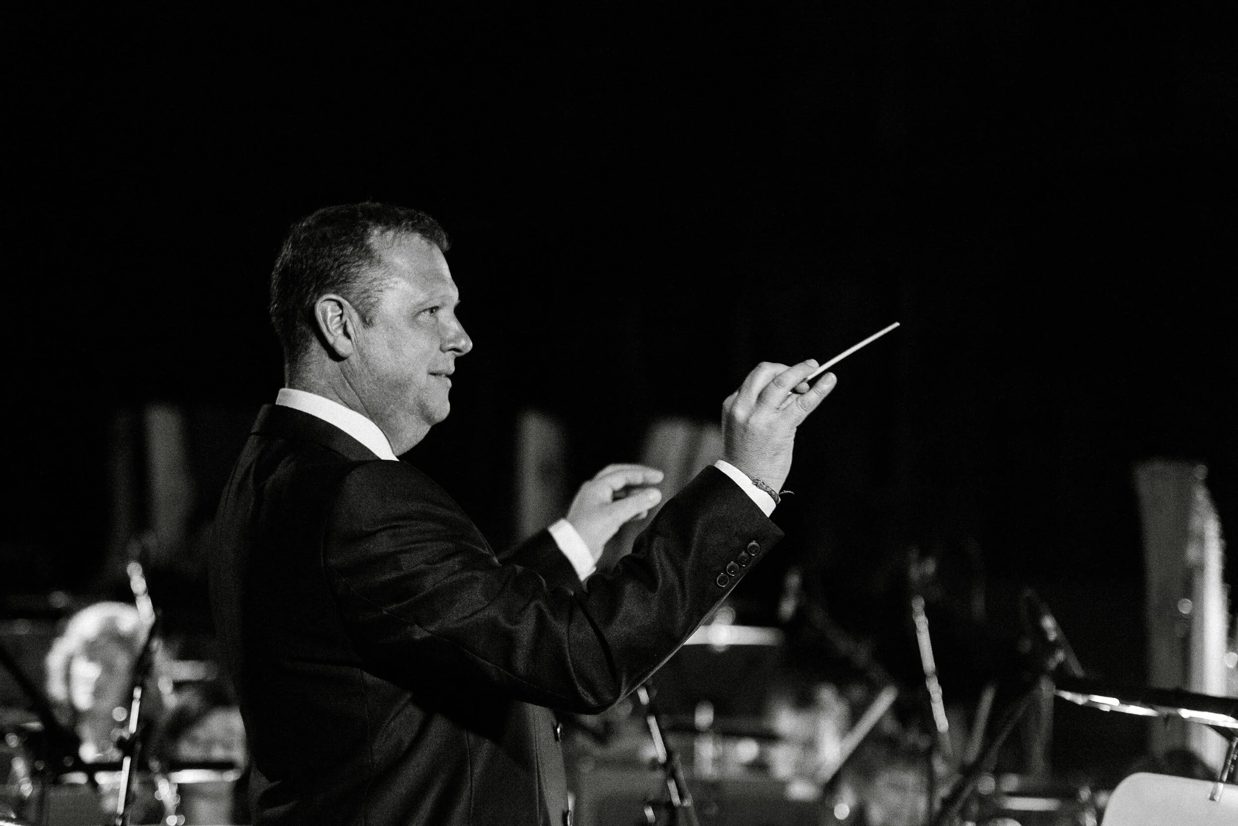 A man in a tuxedo conducts an orchestra with a baton, focusing intently during a performance.