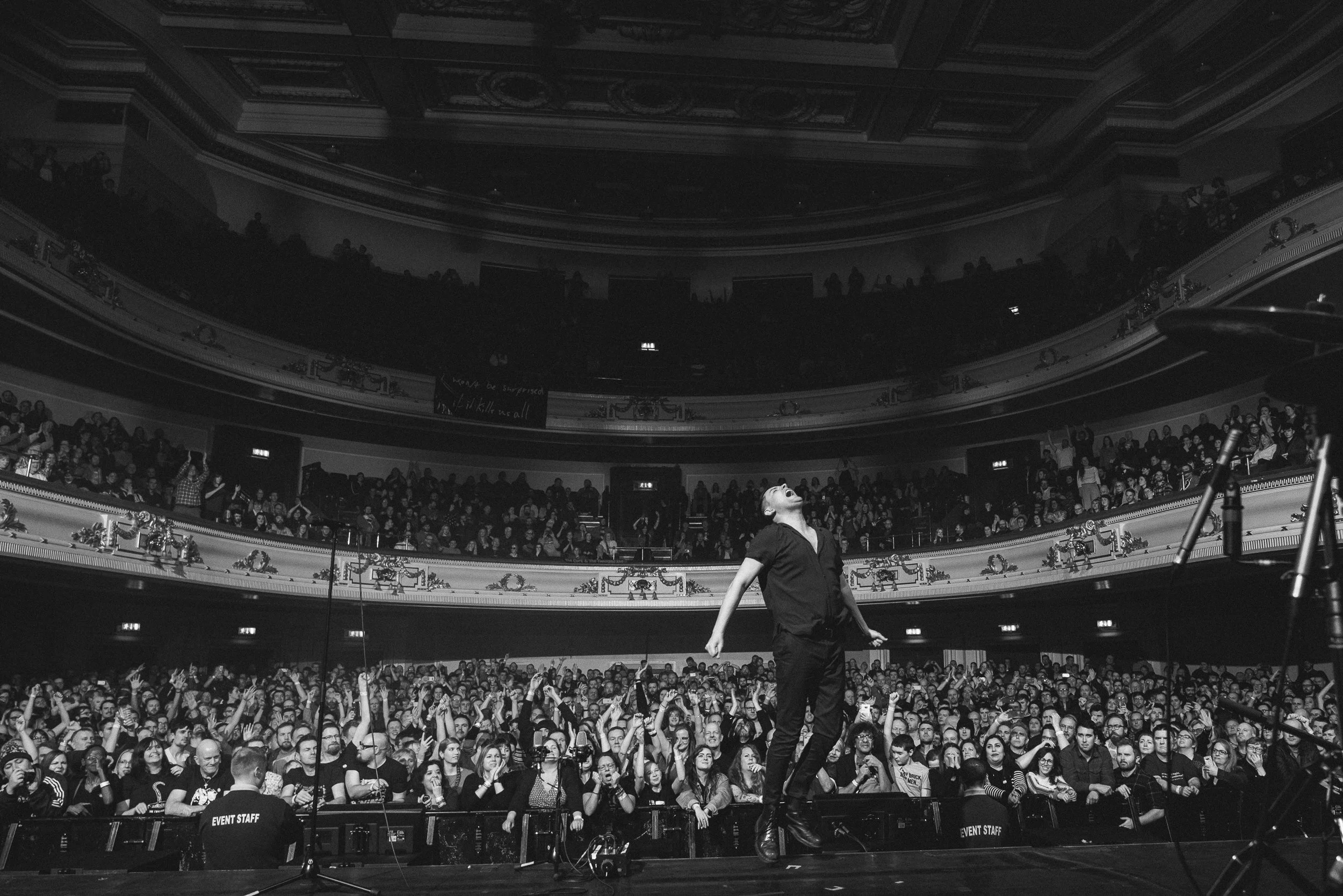 A performer stands center stage in a large theater filled with a seated audience. The theater has multiple levels of balconies decorated with ornate molding and garland. The audience appears engaged as many people raise their hands, and some are taki