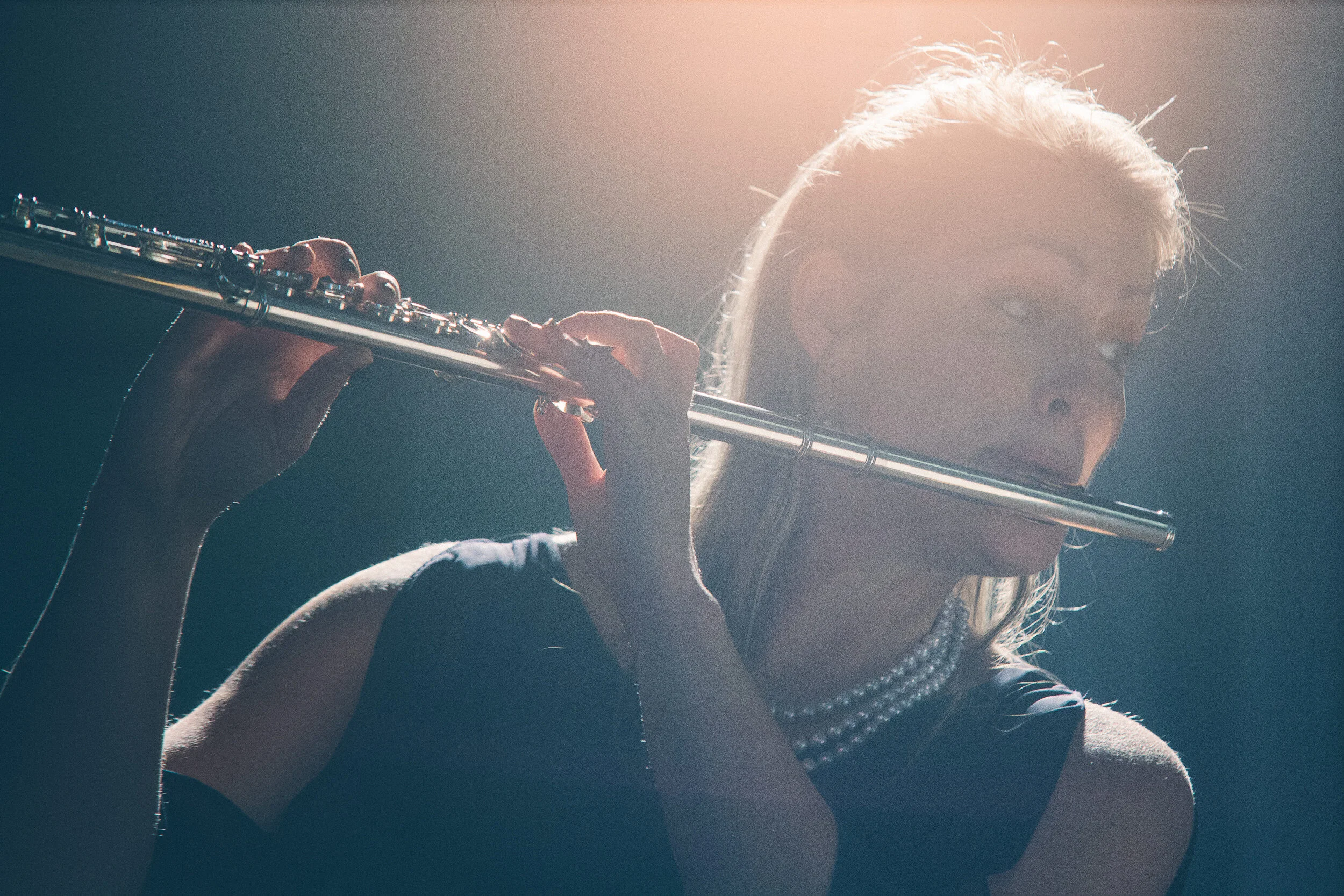 A woman with blonde hair and glasses playing a flute, wearing a pearl necklace and a dark top.