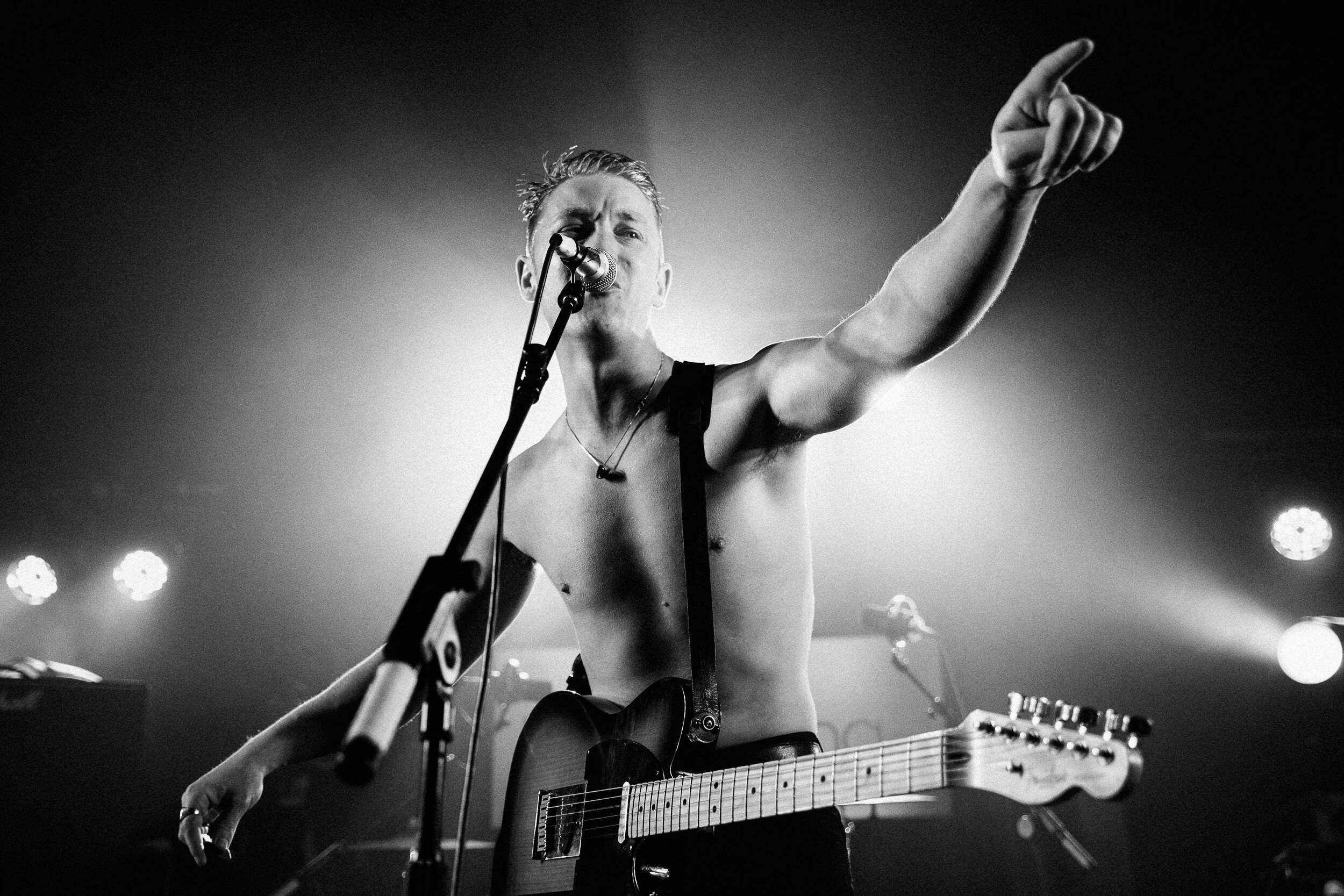 A shirtless male musician with a guitar on stage, singing into a microphone, pointing with his right hand, with stage lights in the background.