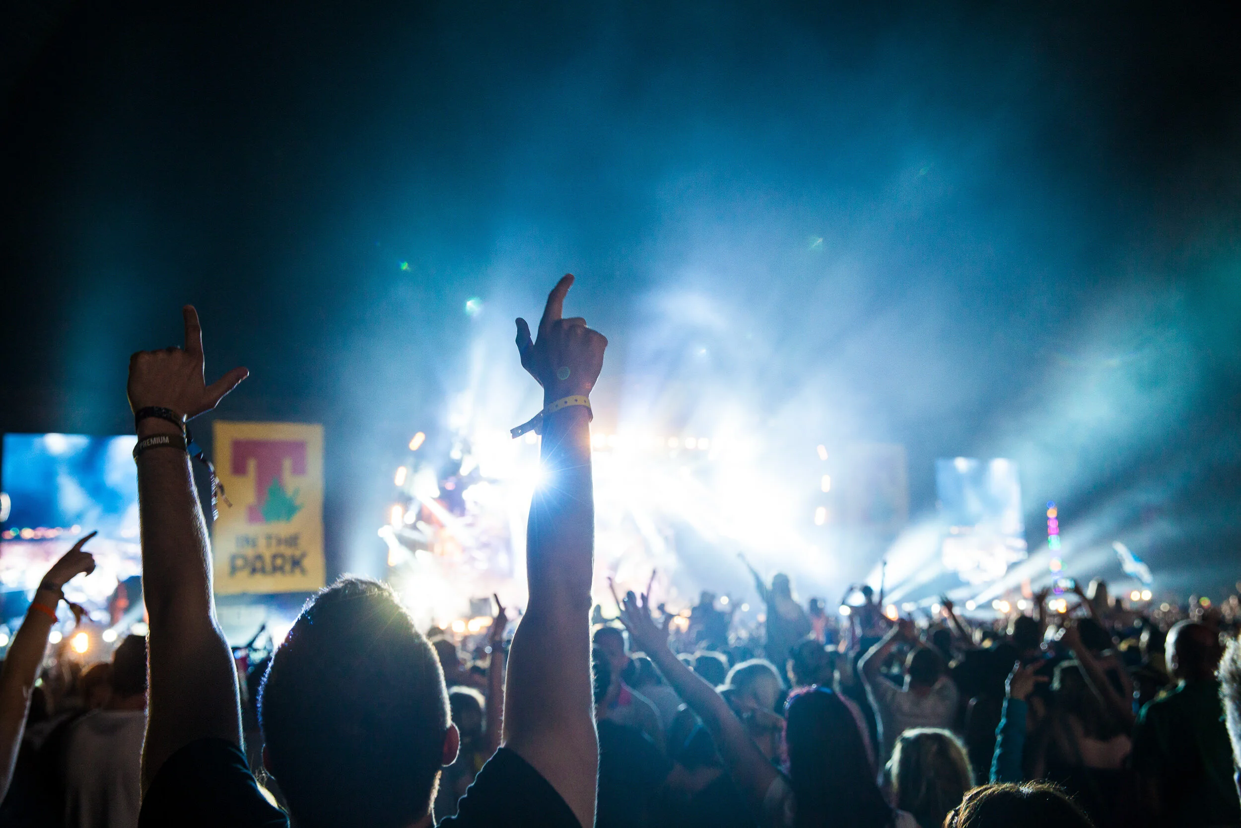Crowd at a concert or music festival with hands raised, illuminated by stage lights and several signs visible in the background.