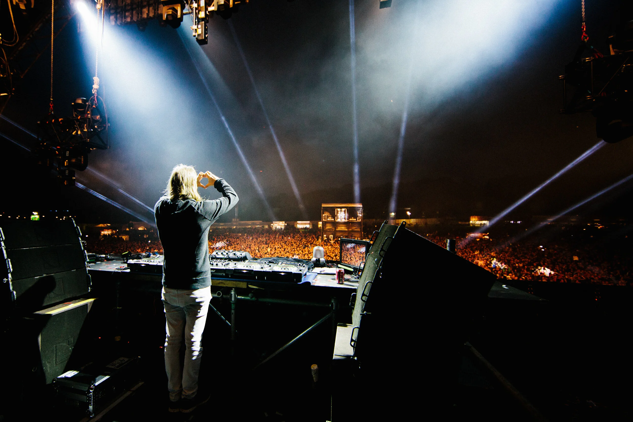 A DJ with long blond hair making a heart shape with their hands in front of a large audience at a concert. The stage is equipped with DJ turntables, mixers, and lighting effects, with bright beams of light shining into the dark night sky.