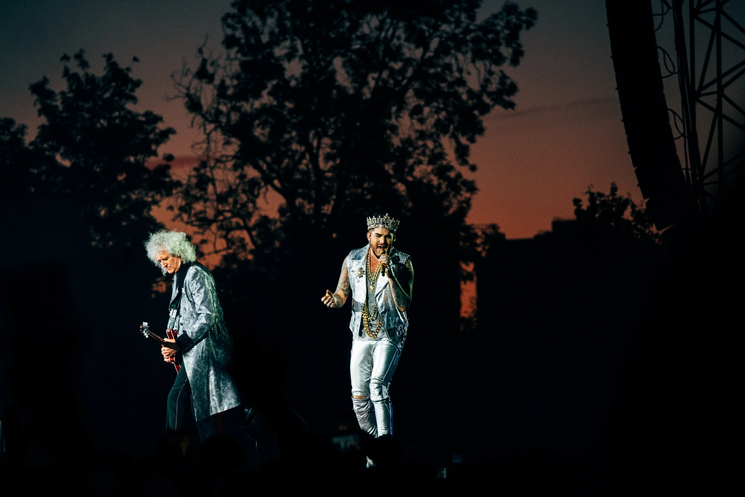 Two male performers on stage at dusk, one with white curly hair playing a guitar and the other with a crown singing into a microphone, both dressed in metallic clothing, with dark silhouettes of trees and a power line in the background.