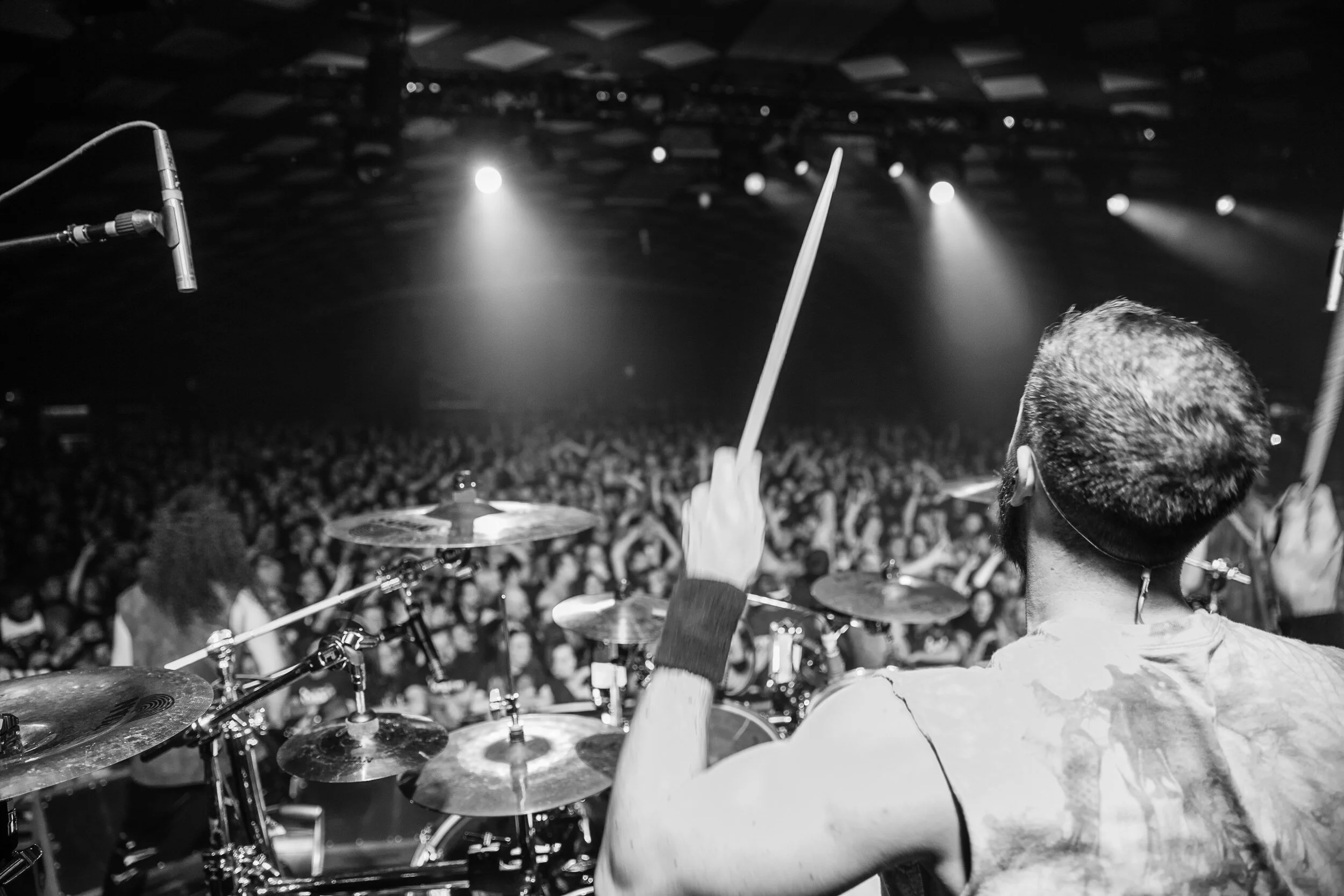 A drummer playing on stage with a large crowd in front, lighting and microphone visible in black and white photo.