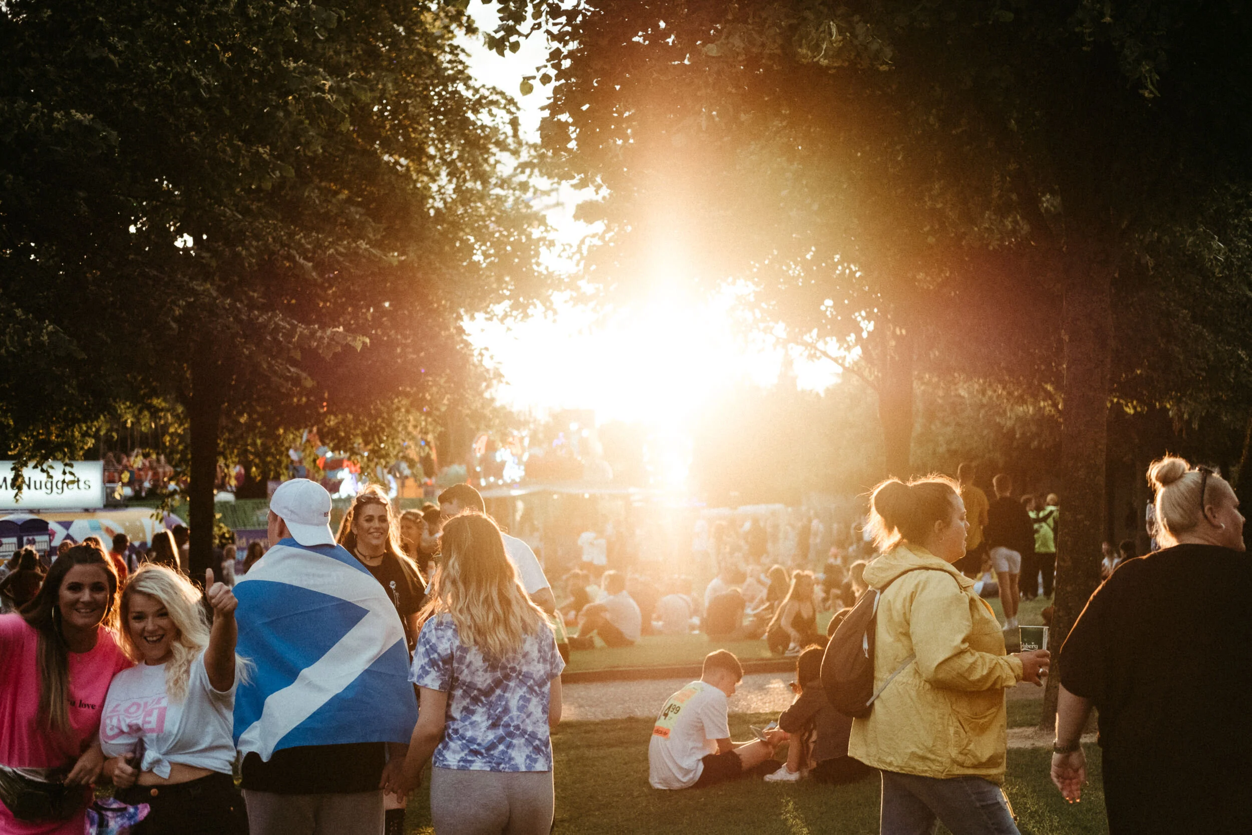 A crowd of people at an outdoor event during sunset, with some people smiling and others engaged in conversation, trees lining the area, and a bright light in the background.