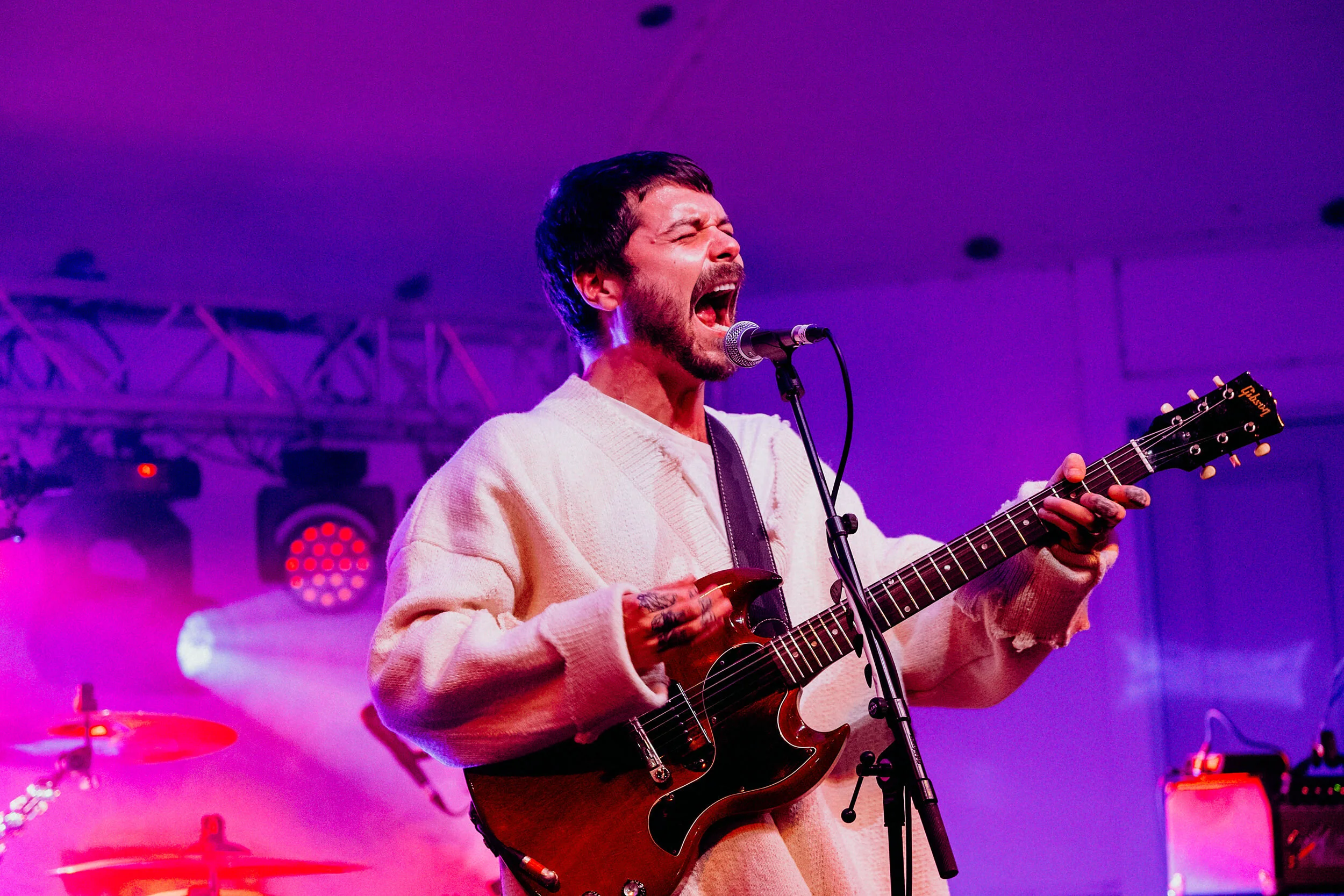 A man singing passionately while playing an electric guitar on stage with purple lighting.